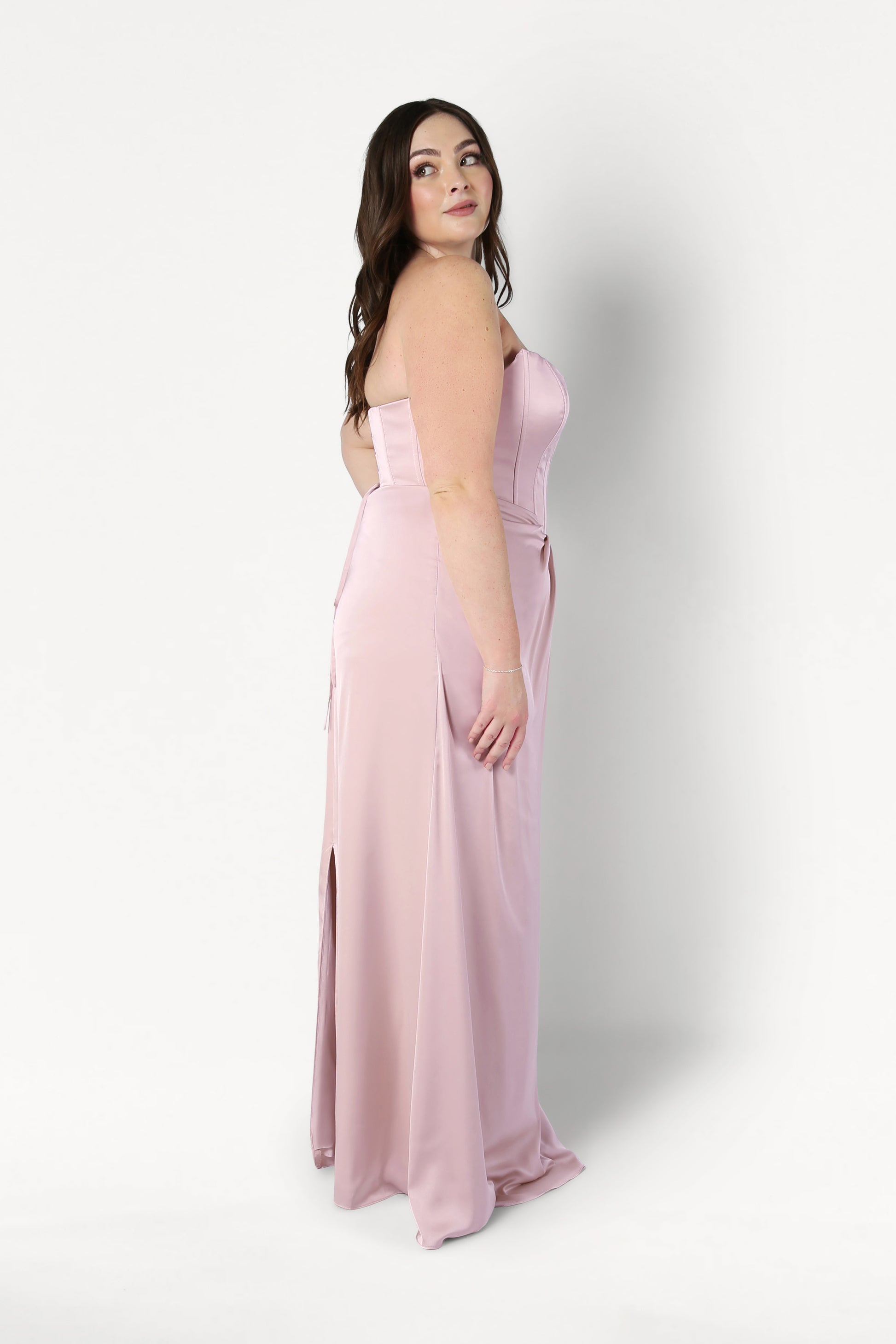 A woman with long brown hair models the size-inclusive Selena Bridesmaid Dress in Blush Pink Satin by TH&TH. The dress features a corset bodice and high slit. She stands in profile against a plain white background, looking over her shoulder.