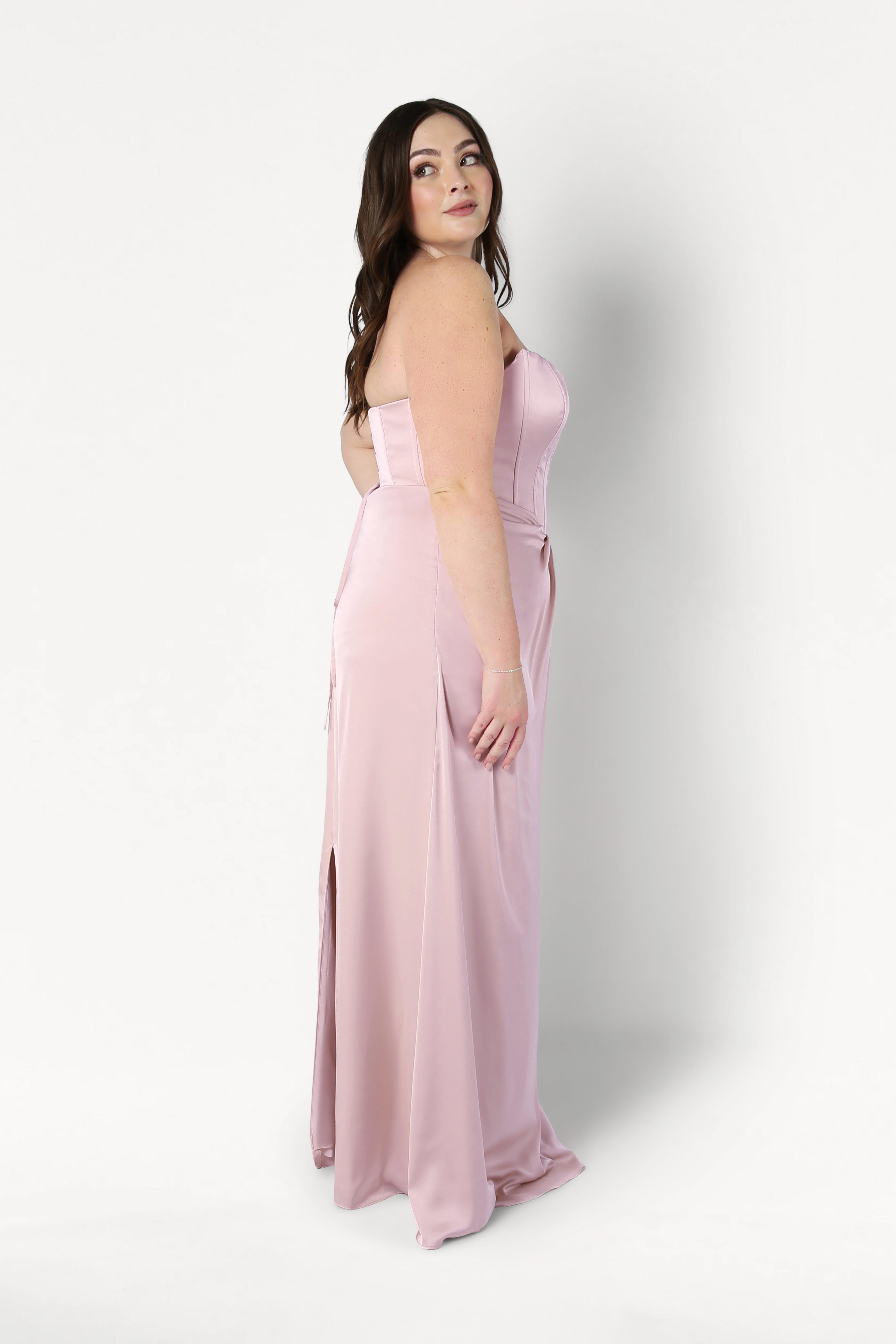 A woman with long brown hair models the size-inclusive Selena Bridesmaid Dress in Blush Pink Satin by TH&TH. The dress features a corset bodice and high slit. She stands in profile against a plain white background, looking over her shoulder.