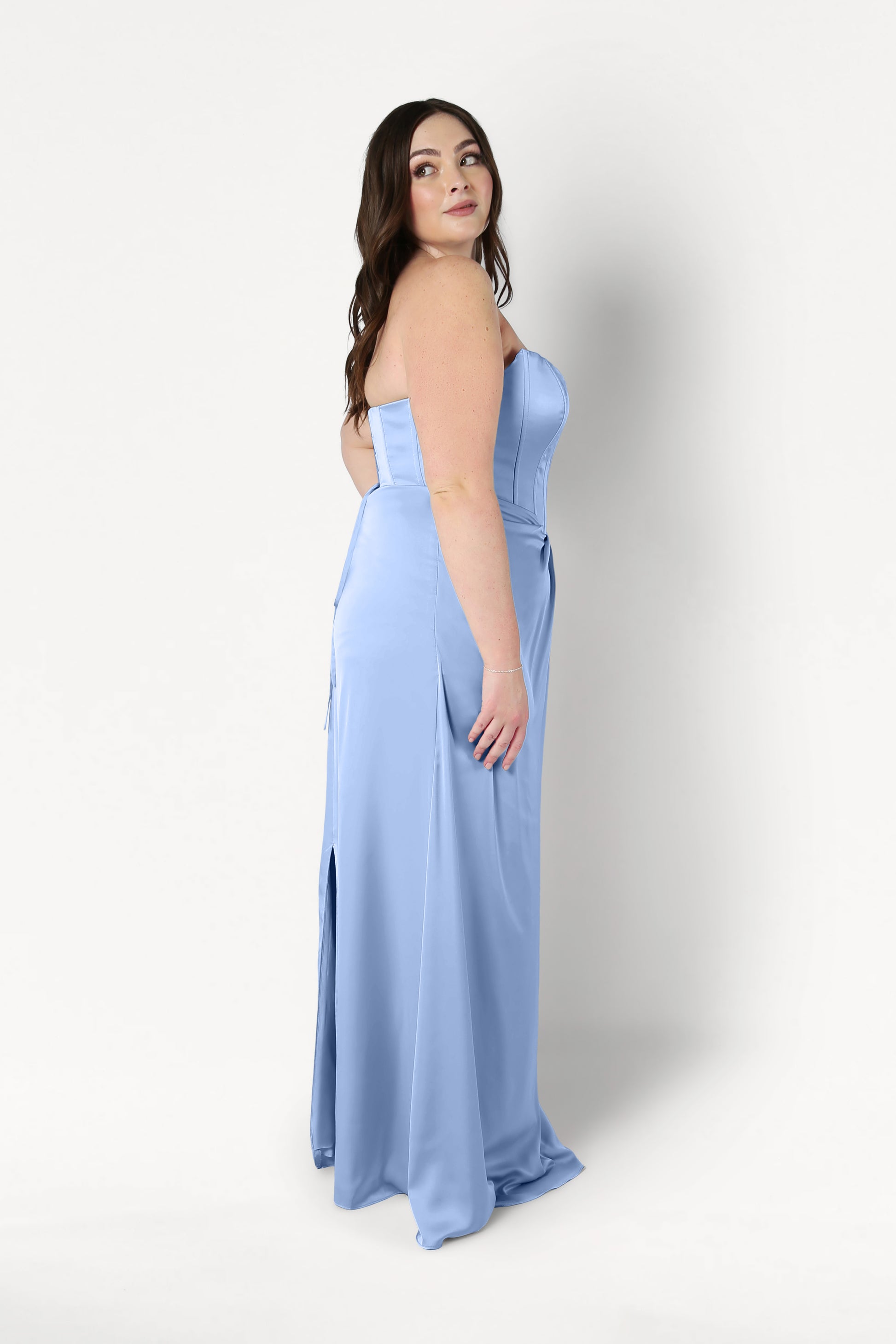 A woman with long brown hair stands in profile against a white background, wearing the TH&TH Selena Bridesmaid Dress in Dusty Blue Satin featuring a strapless corset bodice and front slit.