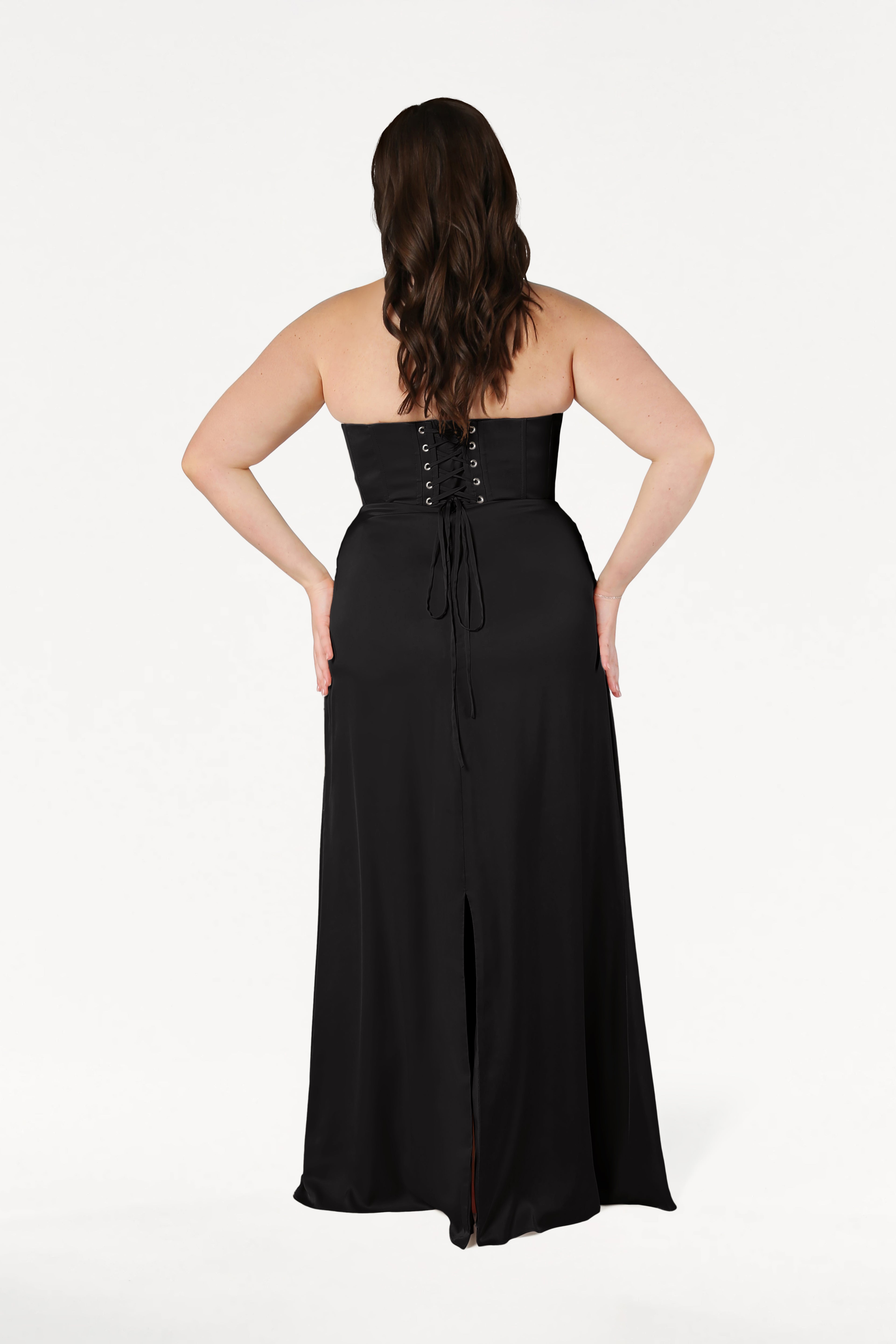 A woman with long wavy brown hair is shown from the back wearing the TH&TH Selena Bridesmaid Dress in Black Satin, which features a strapless lace-up corset bodice and a back slit, against a plain white background.