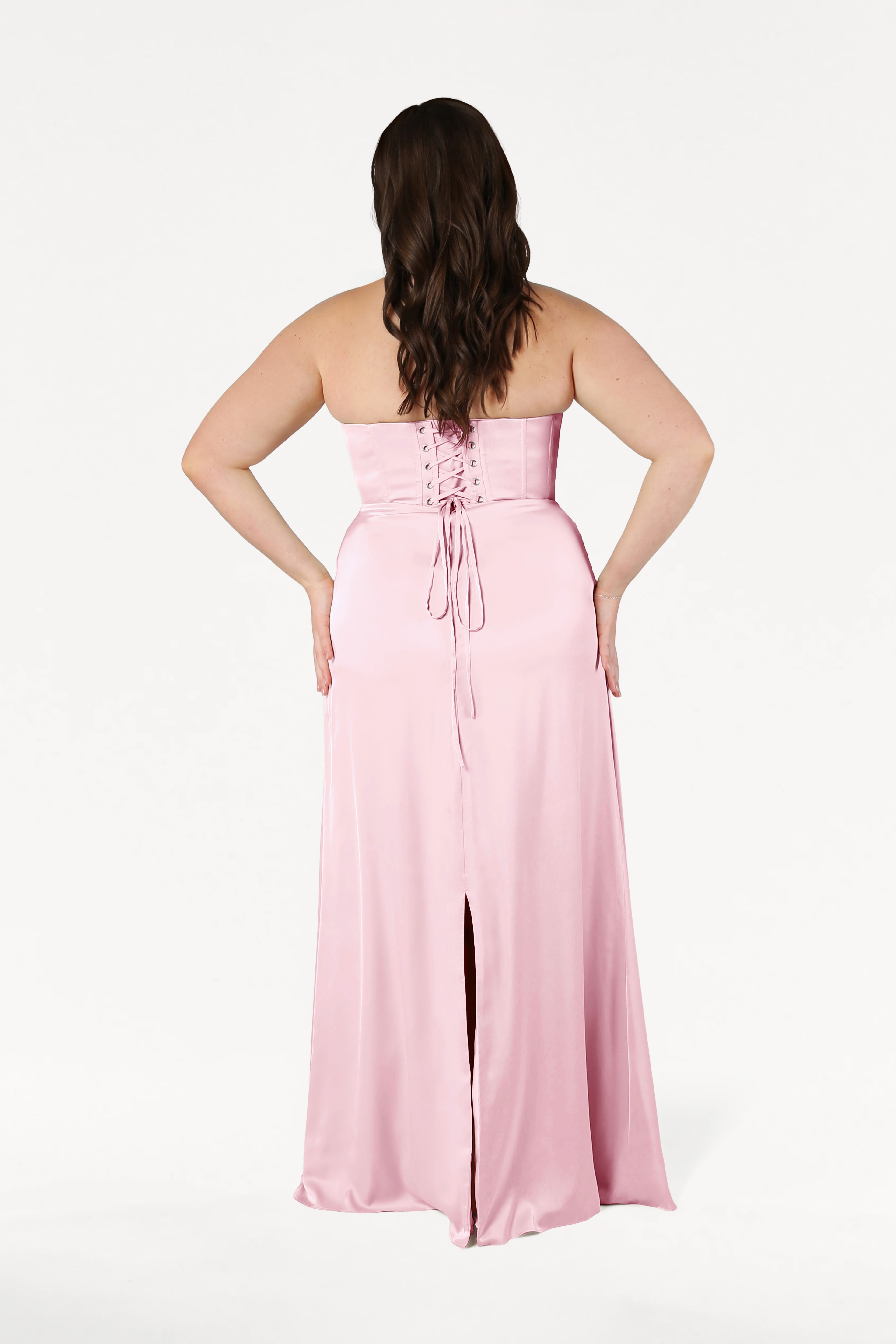 A woman models the TH&TH Selena Bridesmaid Dress in blush pink satin, featuring a strapless lace-up corset back and a center slit, against a plain white background.