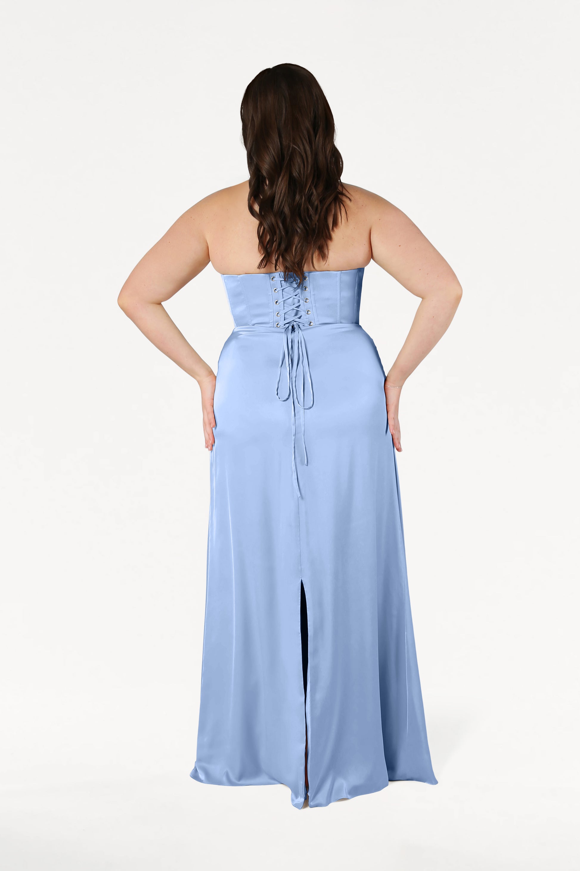 A woman with long brown hair stands facing away, wearing the TH&TH Selena Bridesmaid Dress in Dusty Blue Satin. The size-inclusive gown features a corset bodice, lace-up back, slit skirt, and is set against a plain white background.