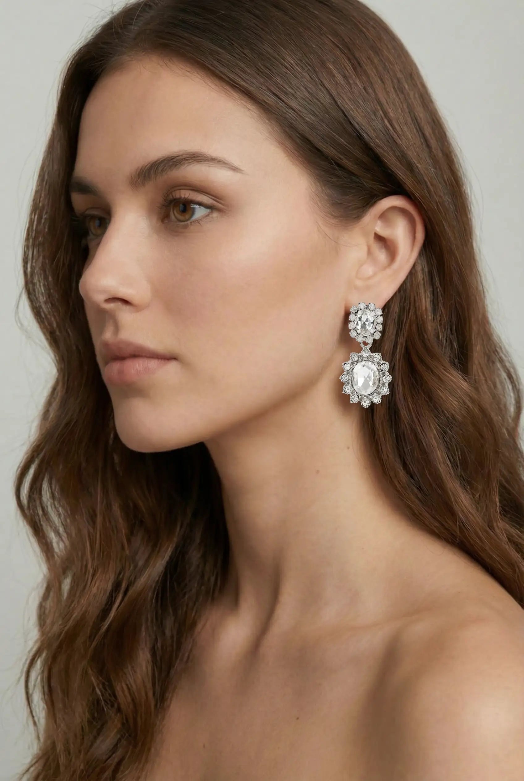 A woman with long, wavy brown hair and bare shoulders is shown in profile, wearing TH&TH’s Imperial Silver Crystal Drop Earrings, which stand out against the plain, neutral background.