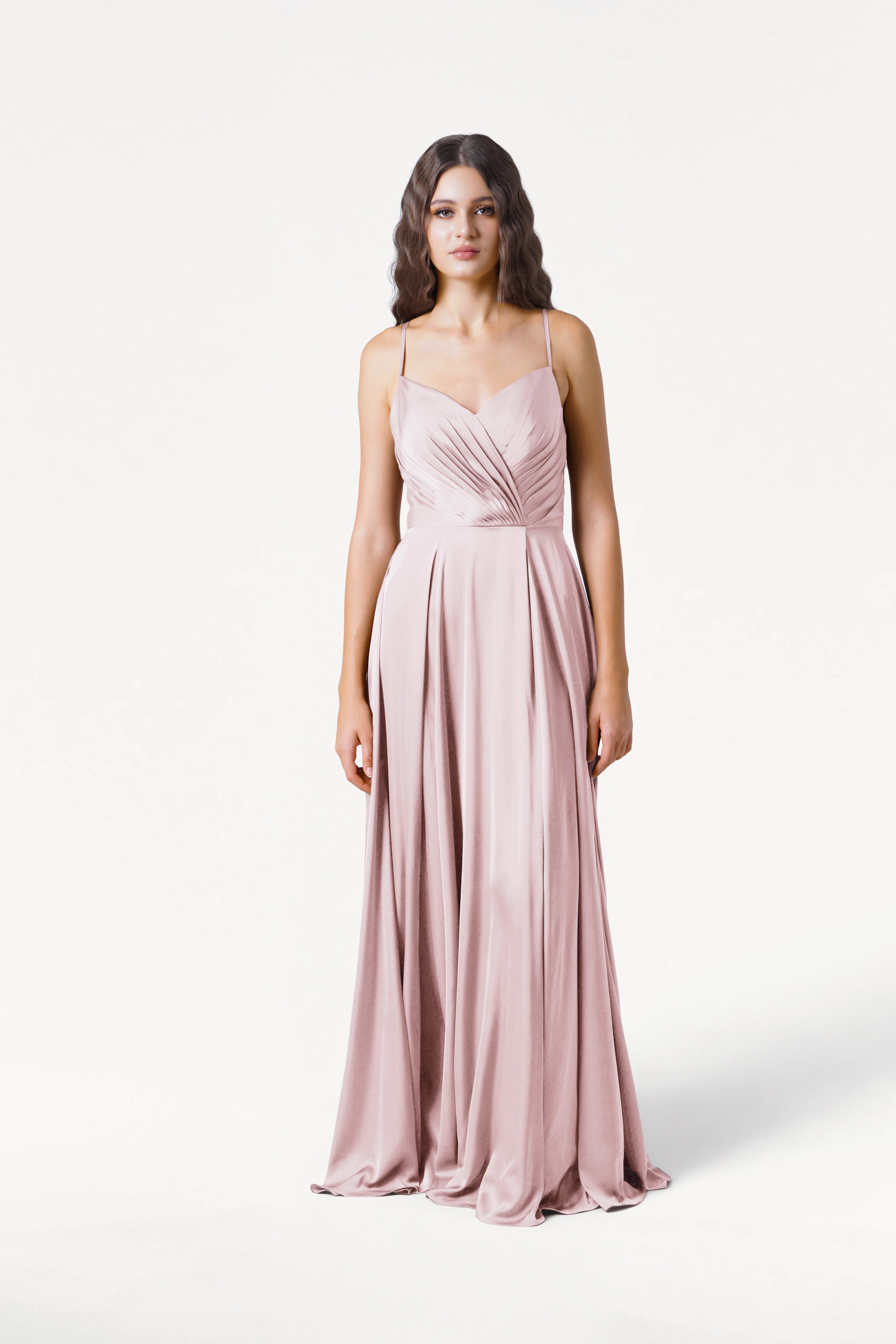 A woman with loose waves stands against a white background wearing the TH&TH Isla Bridesmaid Dress in blush pink satin, a floor-length gown featuring thin straps and a pleated, draped bodice.