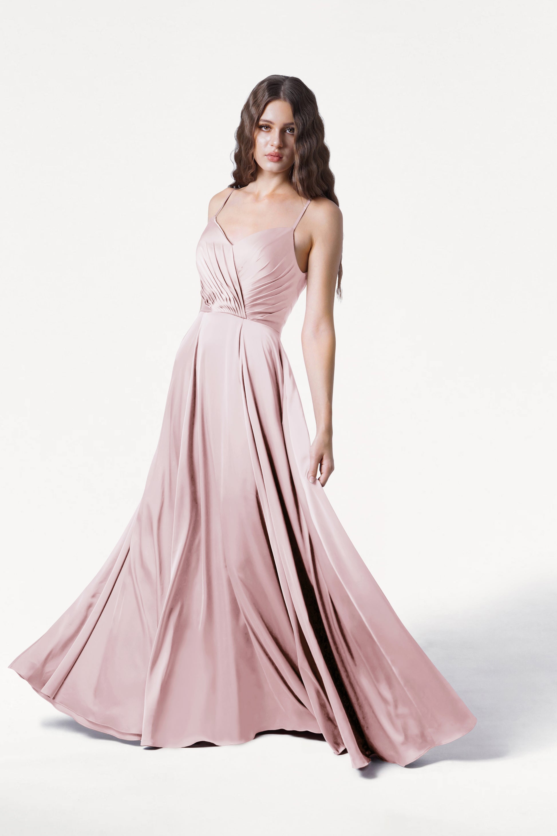 A woman in the TH&TH Isla Bridesmaid Dress in Blush Pink Satin, featuring thin straps and a pleated bodice, stands against a white background, gently holding the skirt as it fans out around her.