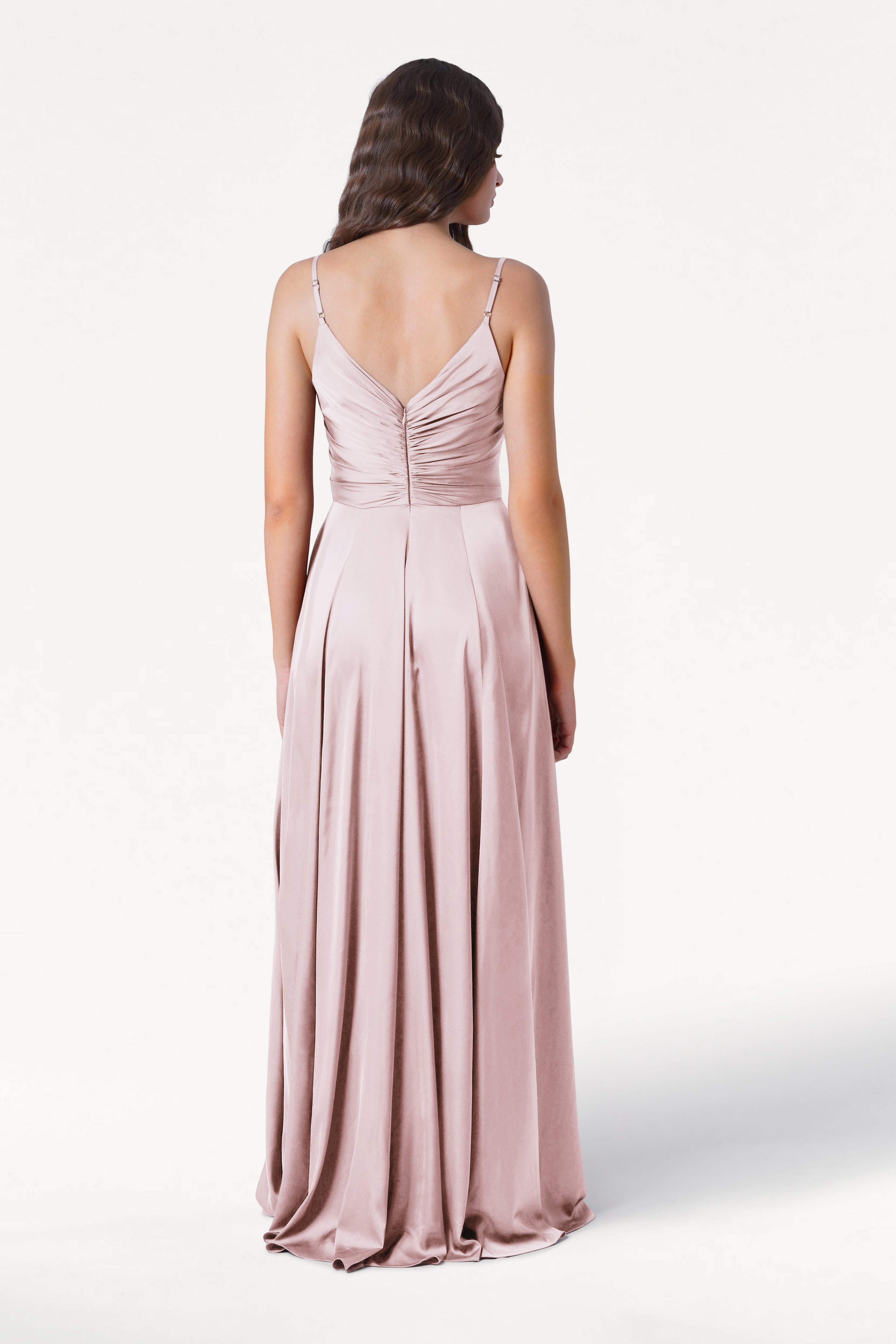A woman with wavy dark hair faces away, wearing the TH&TH Isla Bridesmaid Dress in blush pink satin—an elegant, long gown with thin straps and a pleated bodice, set against a plain white background.