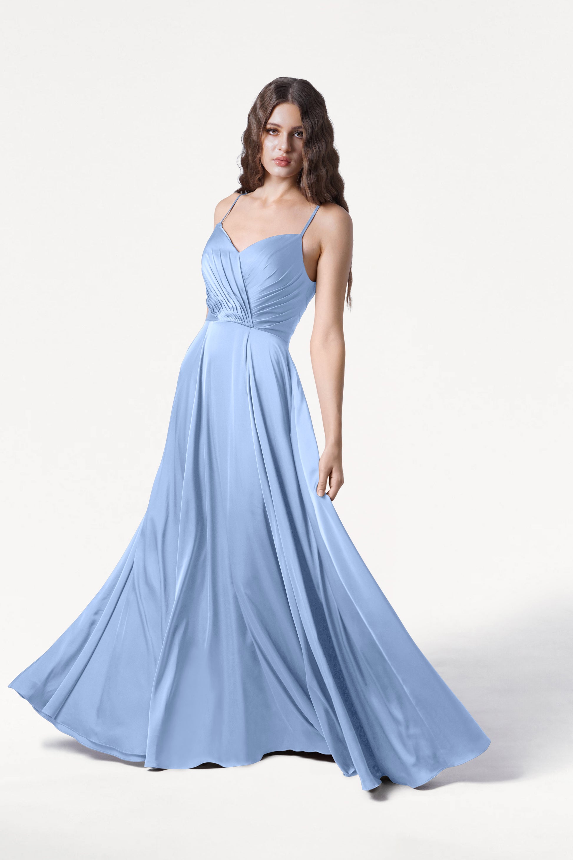 A woman wearing the TH&TH Isla Bridesmaid Dress in Dusty Blue Satin, featuring a pleated bodice and thin straps, stands against a white background, smiling softly with loose wavy hair over her shoulders.