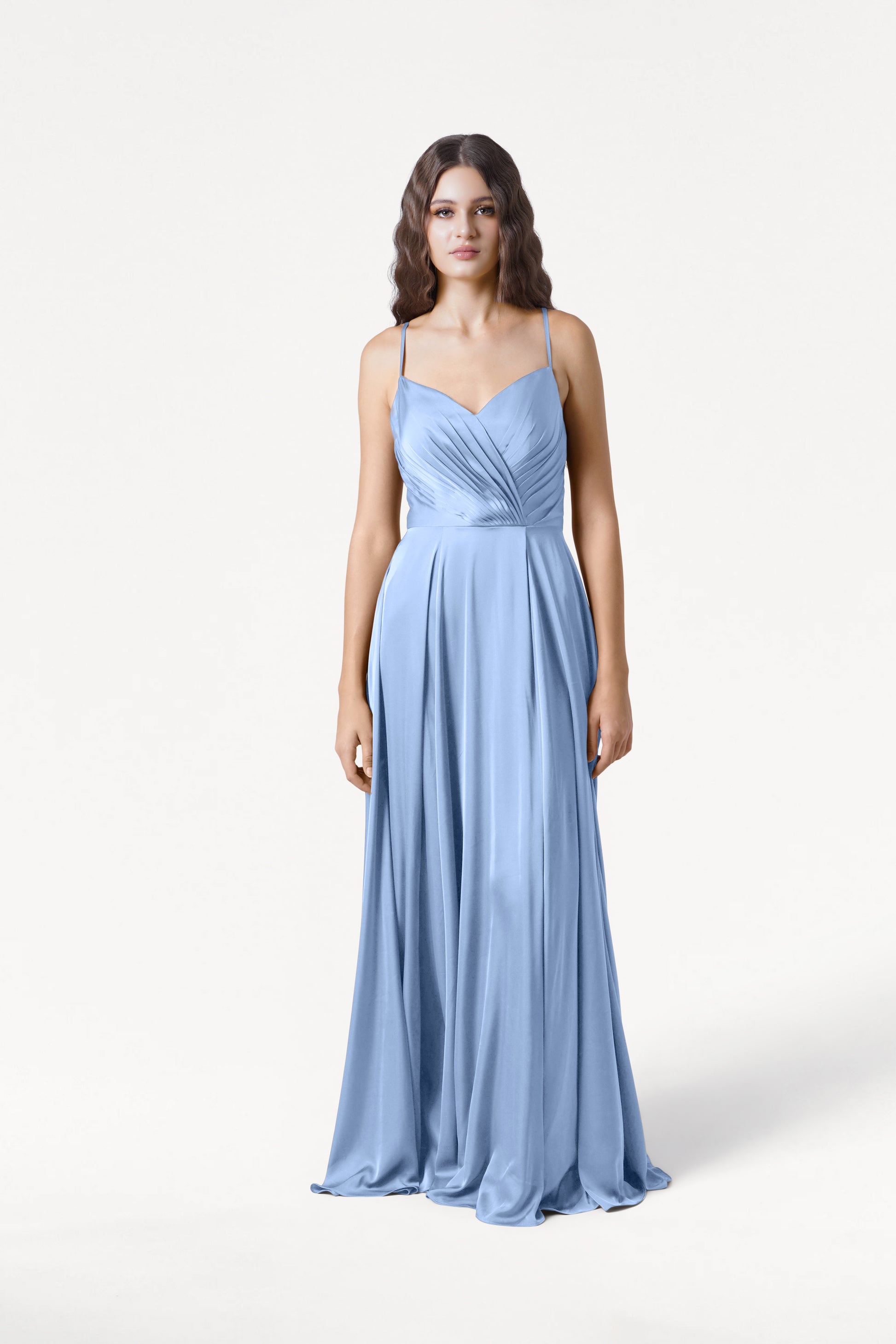 A woman wearing the TH&TH Isla Bridesmaid Dress in Dusty Blue Satin, a long dress with thin straps and a draped, pleated bodice, stands against a plain white background.