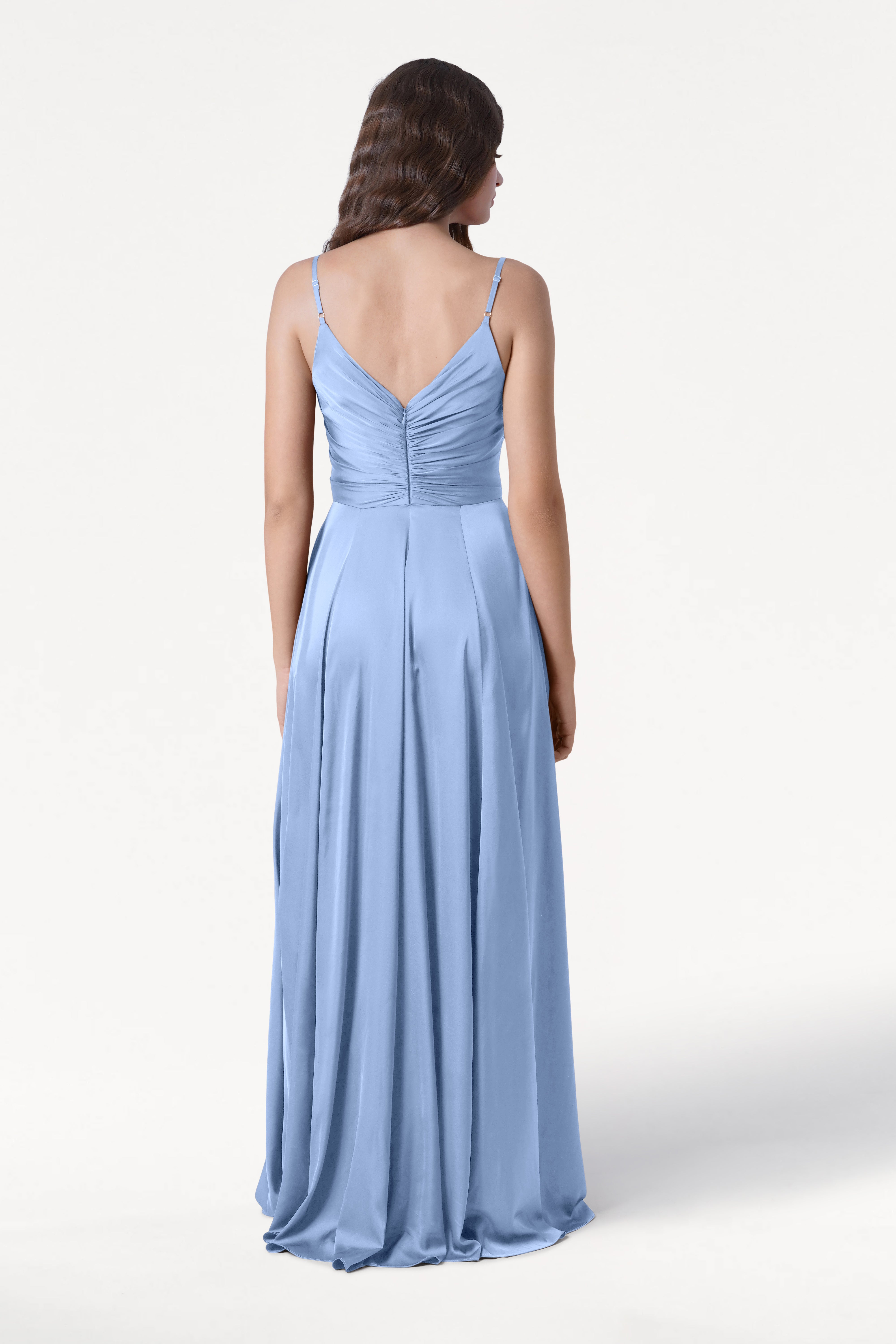 A woman faces away wearing the TH&TH Isla Bridesmaid Dress in Dusty Blue Satin—a floor-length gown with thin straps and a pleated bodice. Her wavy brown hair cascades down her back against a plain white background.