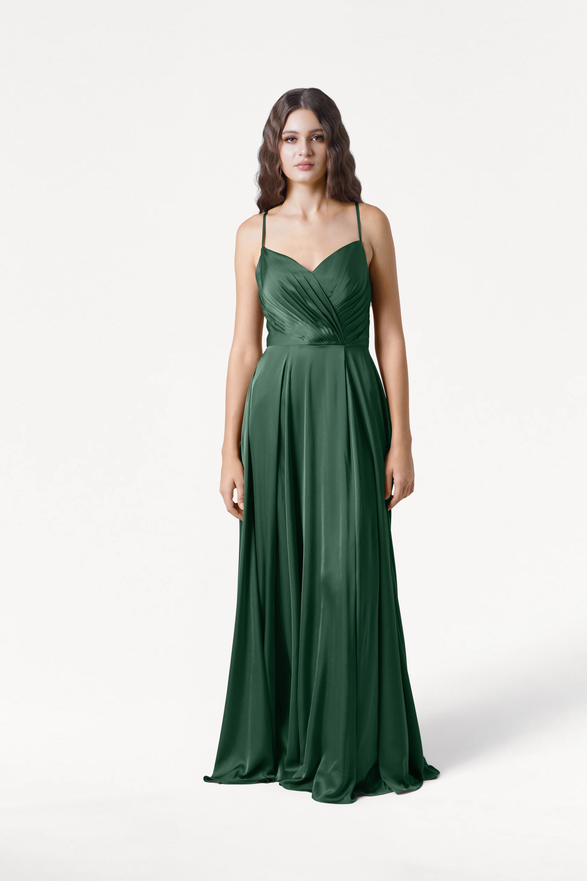 A woman with long, wavy brown hair wears the TH&TH Isla Bridesmaid Dress in Olive Grove Satin, a floor-length dark green dress with thin straps and a fitted, pleated bodice, standing against a plain white background.