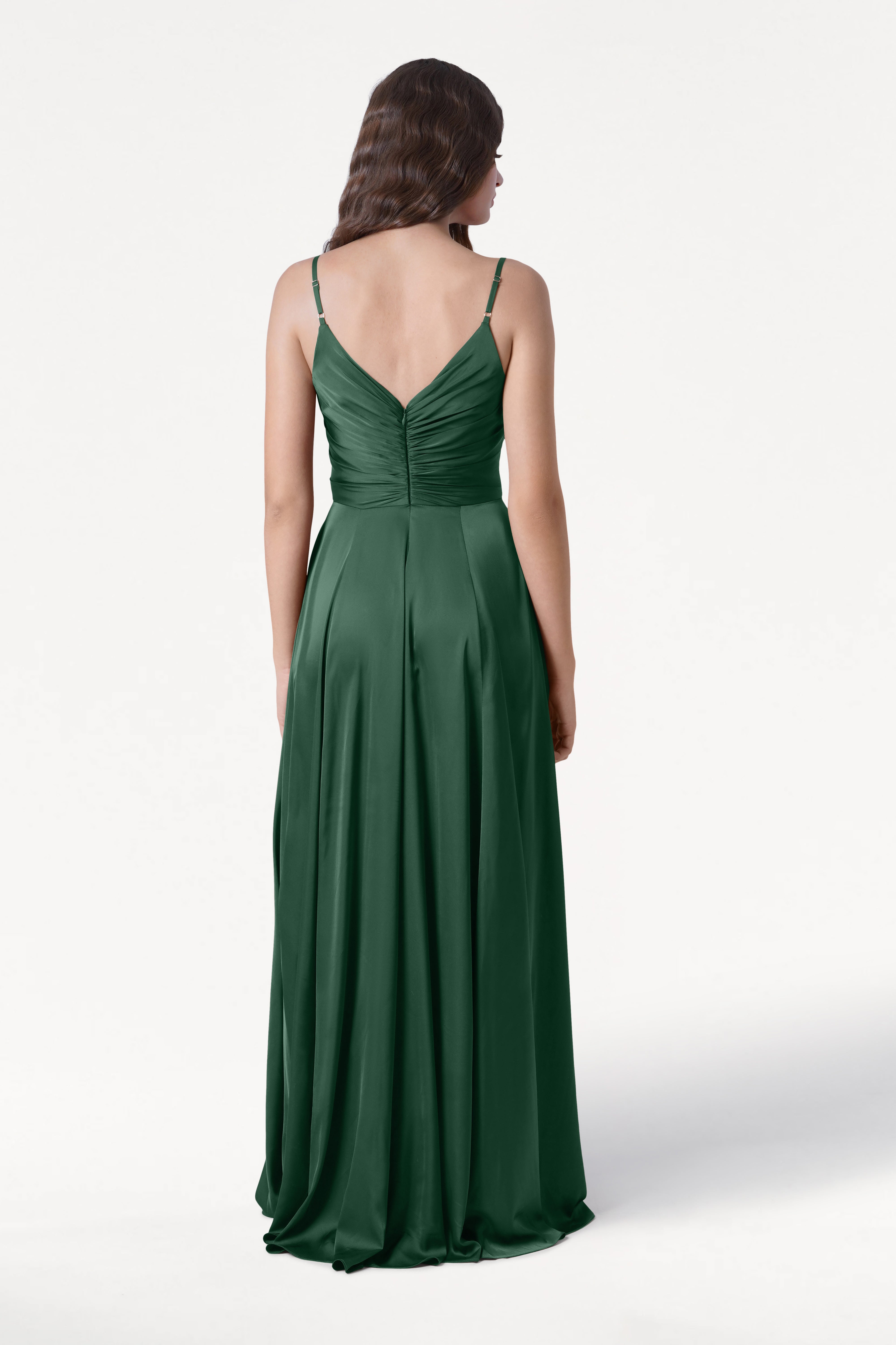 A woman with wavy brown hair models the TH&TH Isla Bridesmaid Dress in Olive Grove Satin, a long dark green pleated gown with thin straps and a V-shaped back, standing against a plain white background, facing away from the camera.