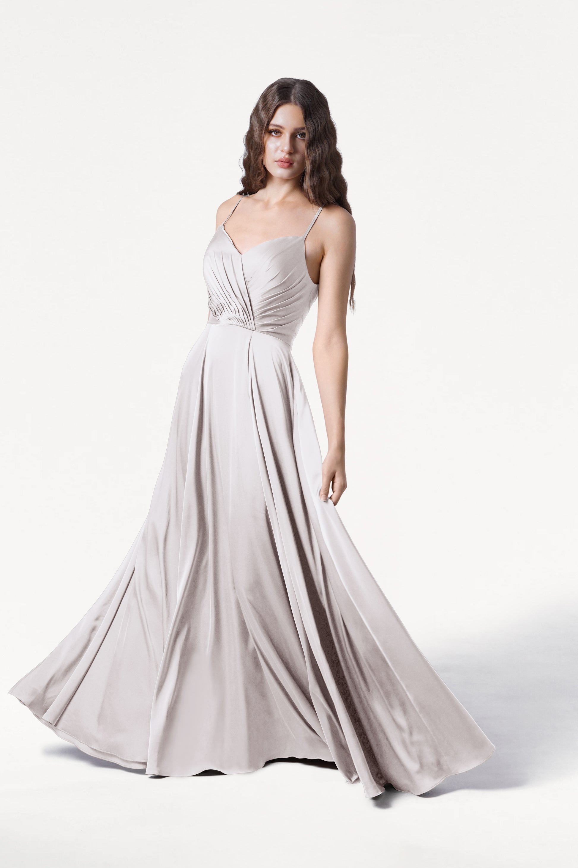 A woman with wavy brown hair stands against a white background wearing the Isla Bridesmaid Dress in Oyster Satin by TH&TH—a long, light gray satin gown with thin straps, a fitted pleated bodice, and an elegant floor-length skirt.