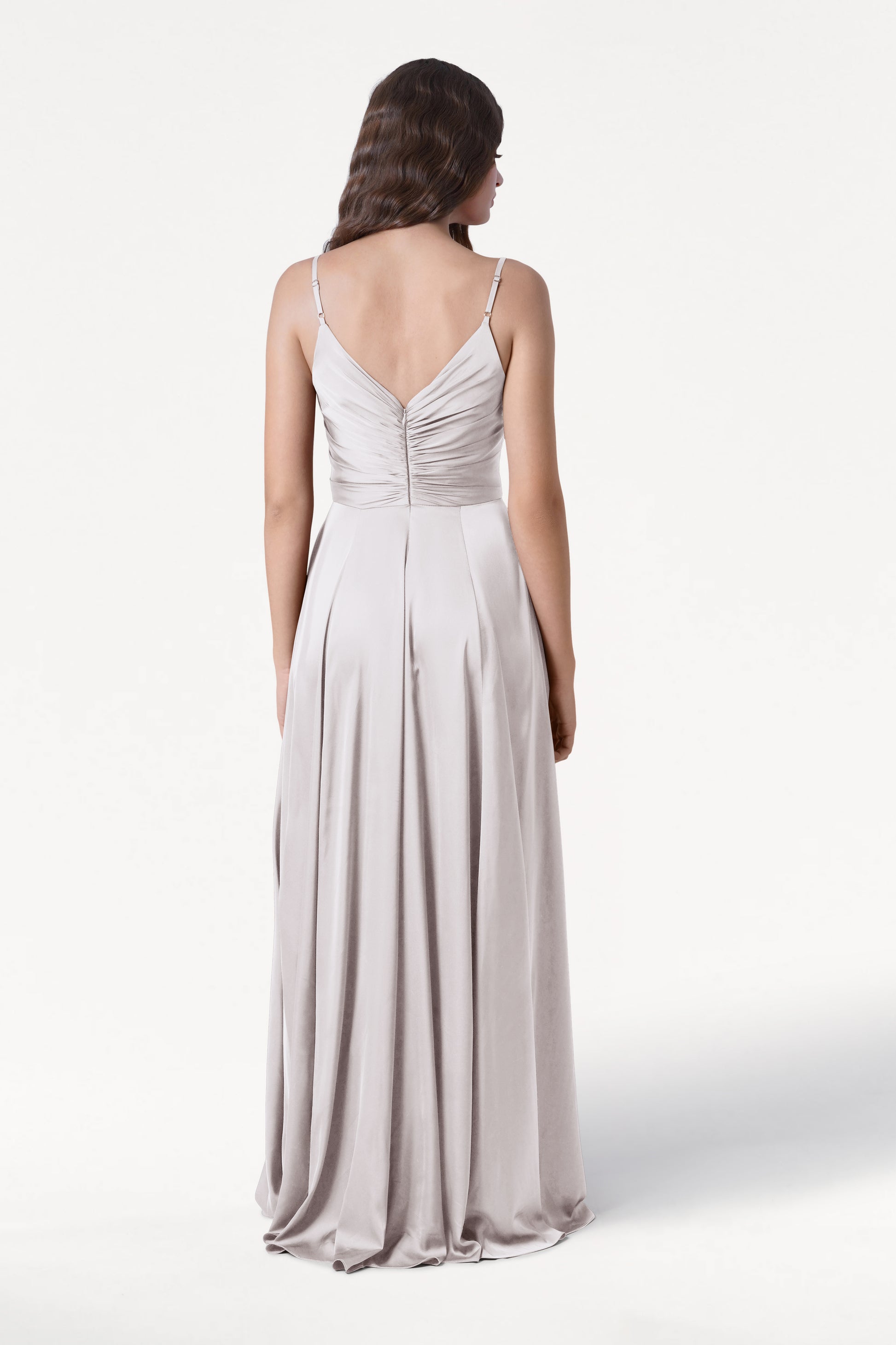 A woman with wavy brown hair stands facing away, wearing the TH&TH Isla Bridesmaid Dress in Oyster Satin—a long, light gray satin gown with a pleated bodice, thin straps, ruched V-back, and flowing skirt against a plain white background.