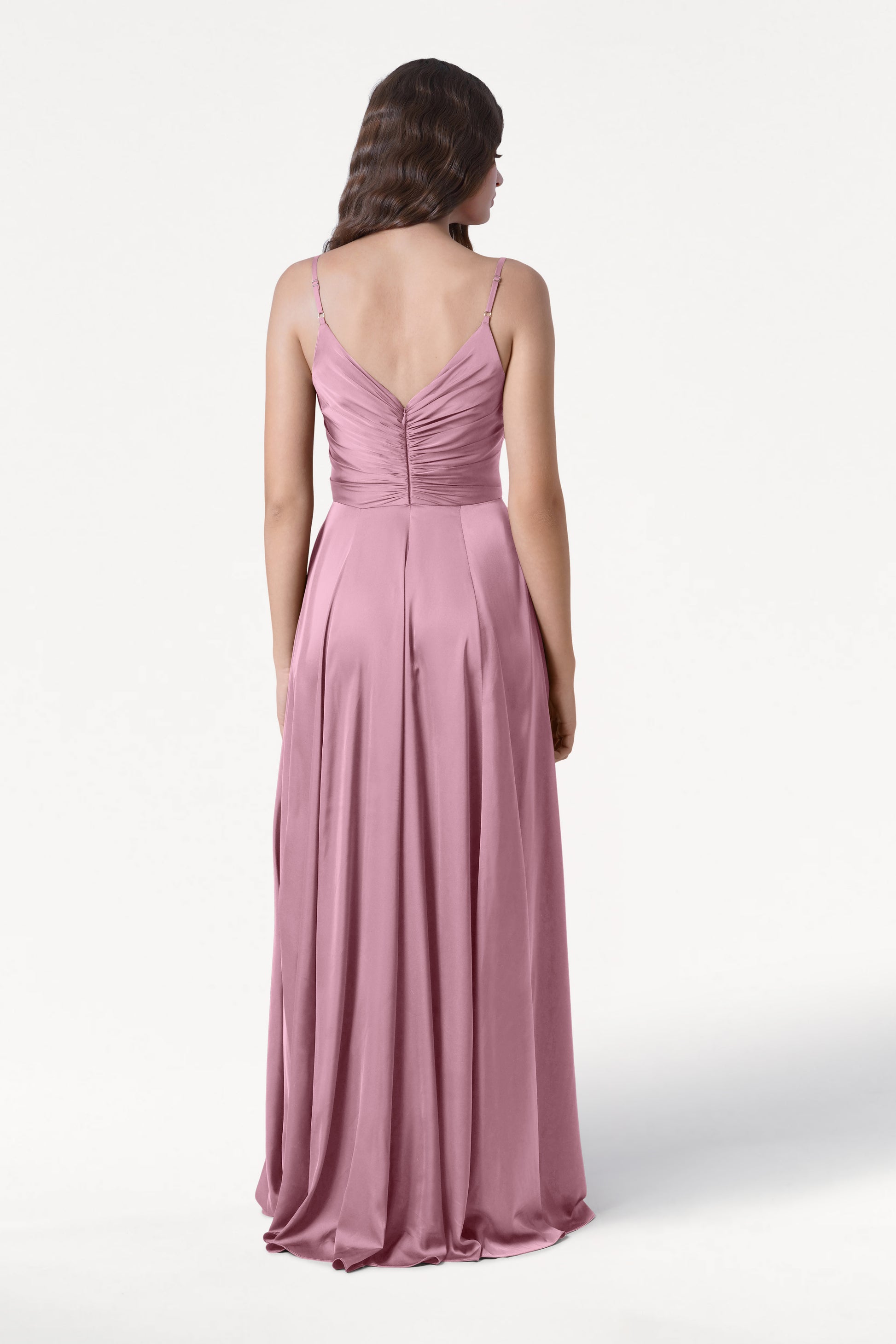 A woman with long, wavy hair faces away, wearing the TH&TH Isla Bridesmaid Dress in rose pink satin featuring adjustable straps and a ruched bodice, set against a plain white background.