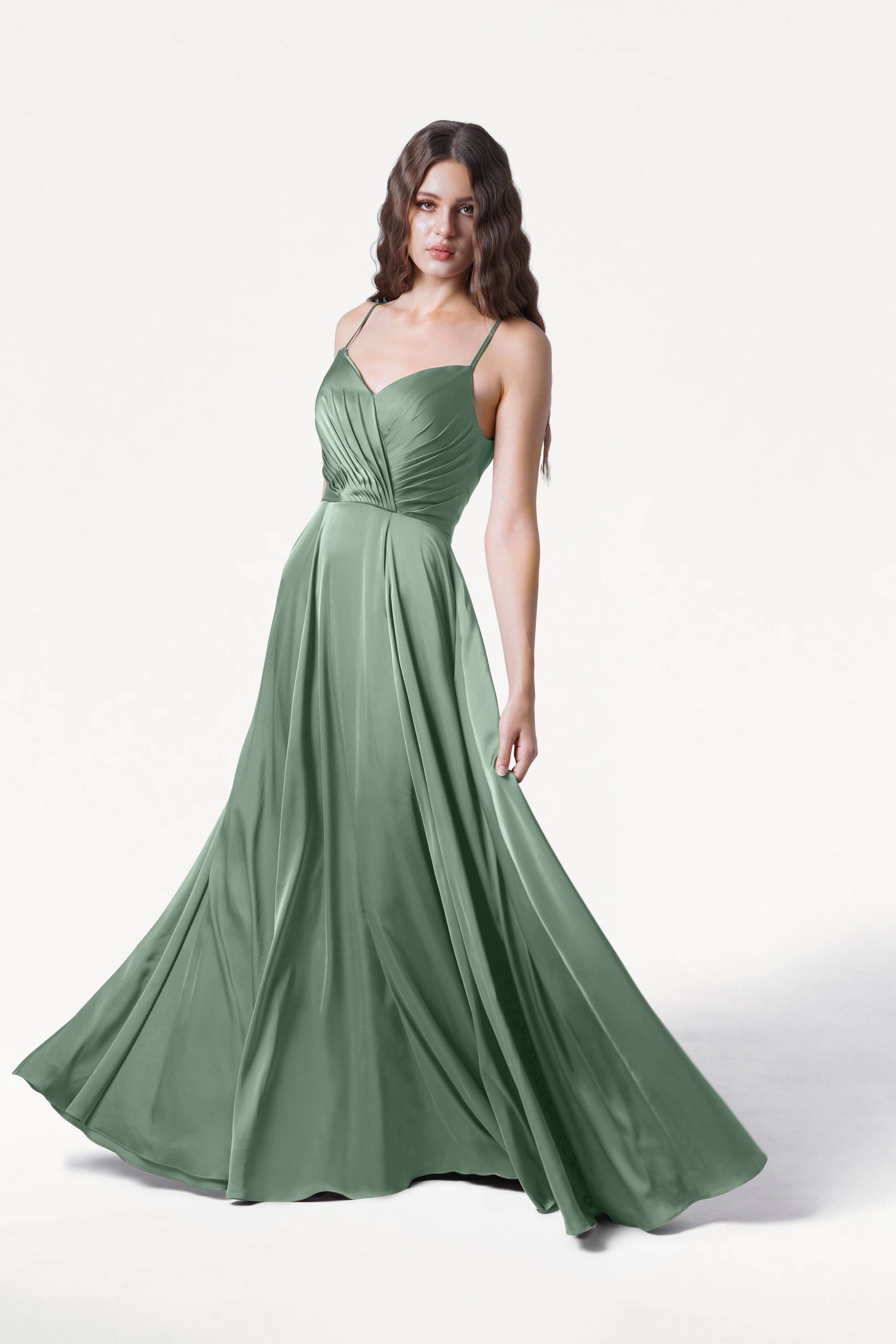 A woman stands against a plain white background wearing the TH&TH Isla Bridesmaid Dress in sage green satin, featuring thin straps, a fitted pleated bodice, and a flowing floor-length flared skirt.