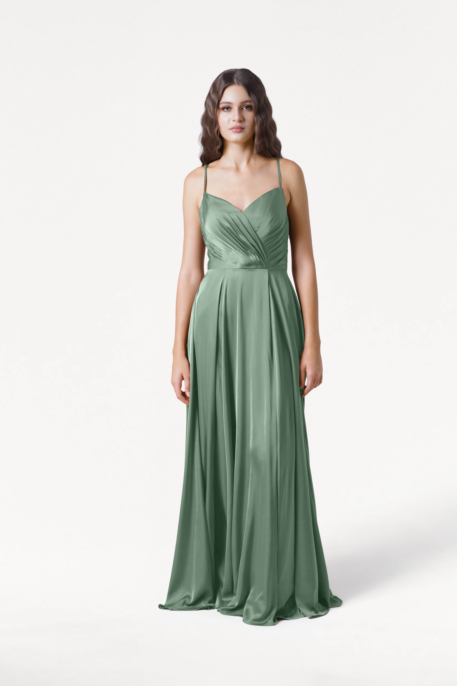 A woman with long, wavy dark hair stands against a white background wearing the TH&TH Isla Bridesmaid Dress in sage green satin—a floor-length, sleeveless gown with a fitted pleated bodice, V-neckline, and flowing skirt.