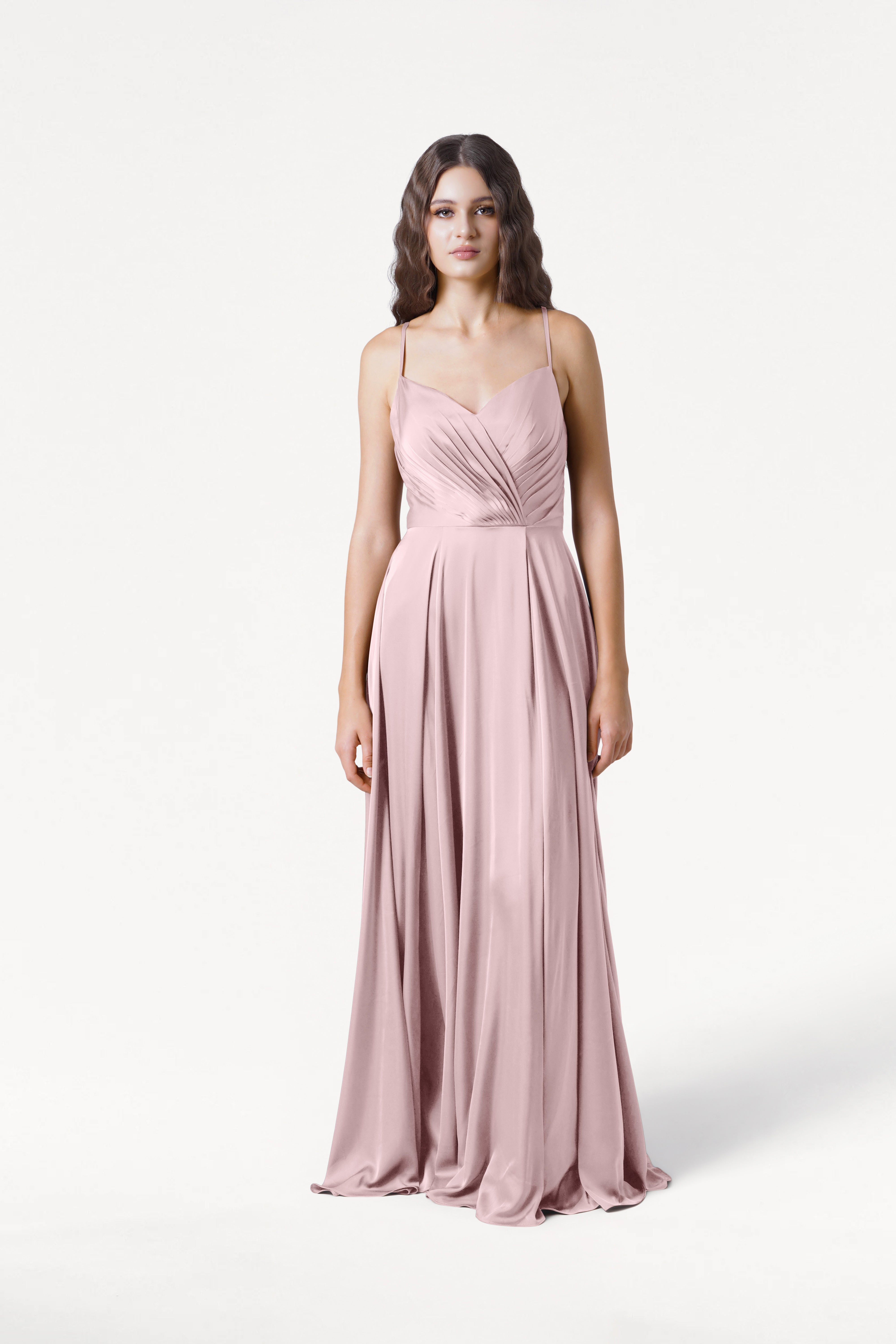 A woman with long, wavy brown hair and a neutral expression stands against a white background wearing the TH&TH Isla Bridesmaid Dress in Smoked Blush Satin, a long gown with thin straps and a draped, pleated bodice.
