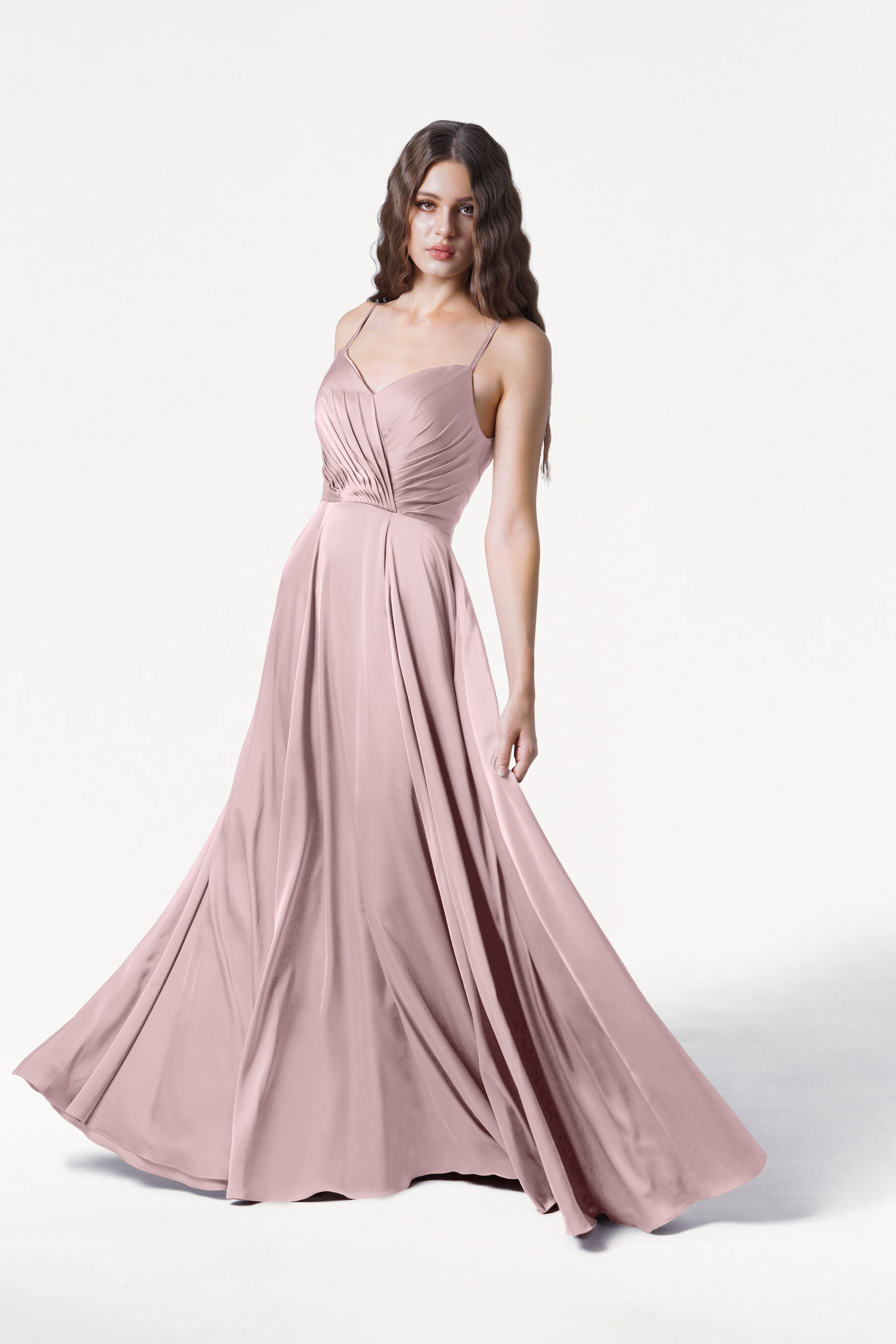 A woman in the TH&TH Isla Bridesmaid Dress in Smoked Blush Satin, featuring a pleated bodice, thin straps, and a flowing floor-length skirt, stands against a plain white background.