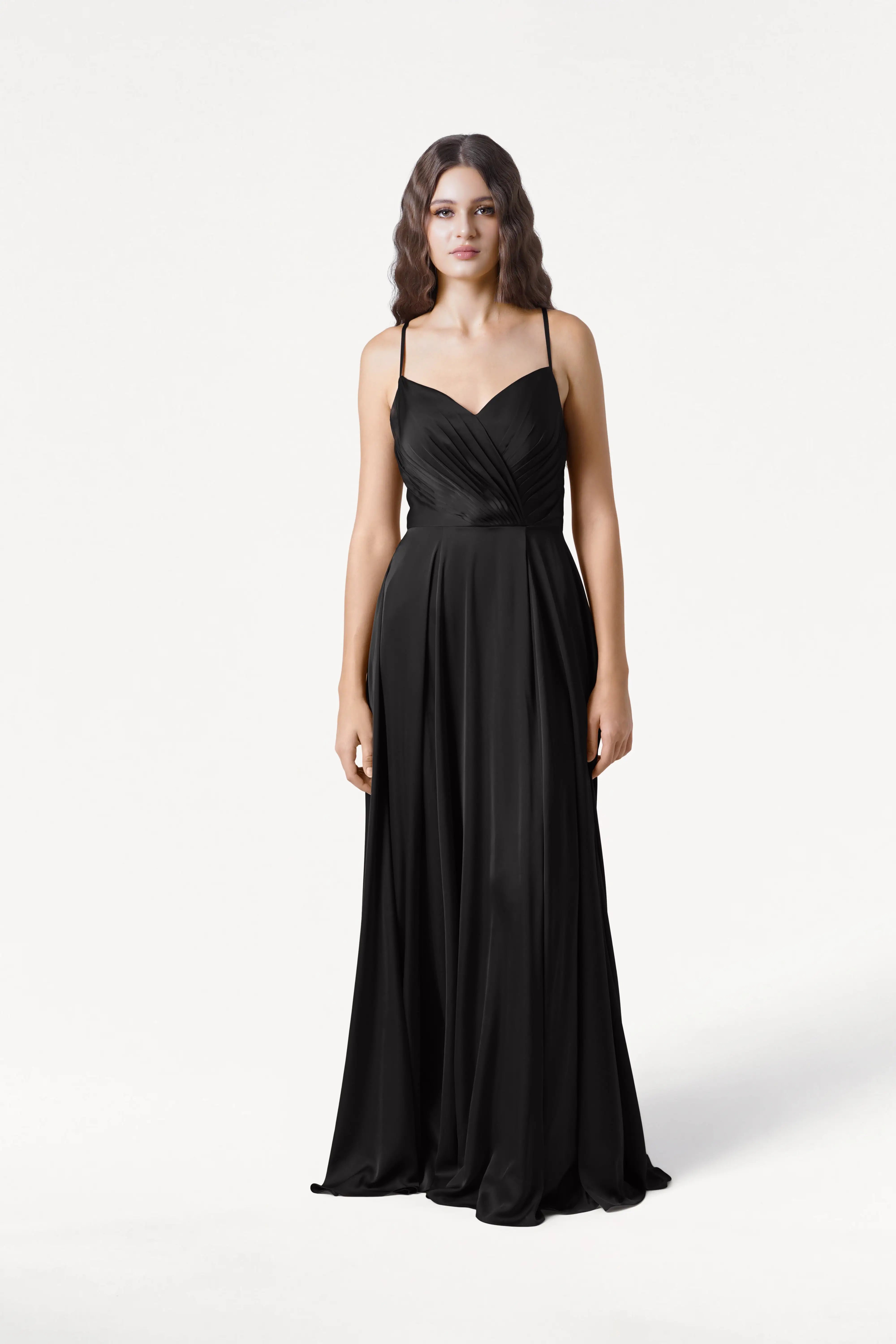 A woman with loose waves and a neutral expression stands against a white background, wearing the TH&TH Isla Bridesmaid Dress in black satin featuring thin straps and a pleated bodice.