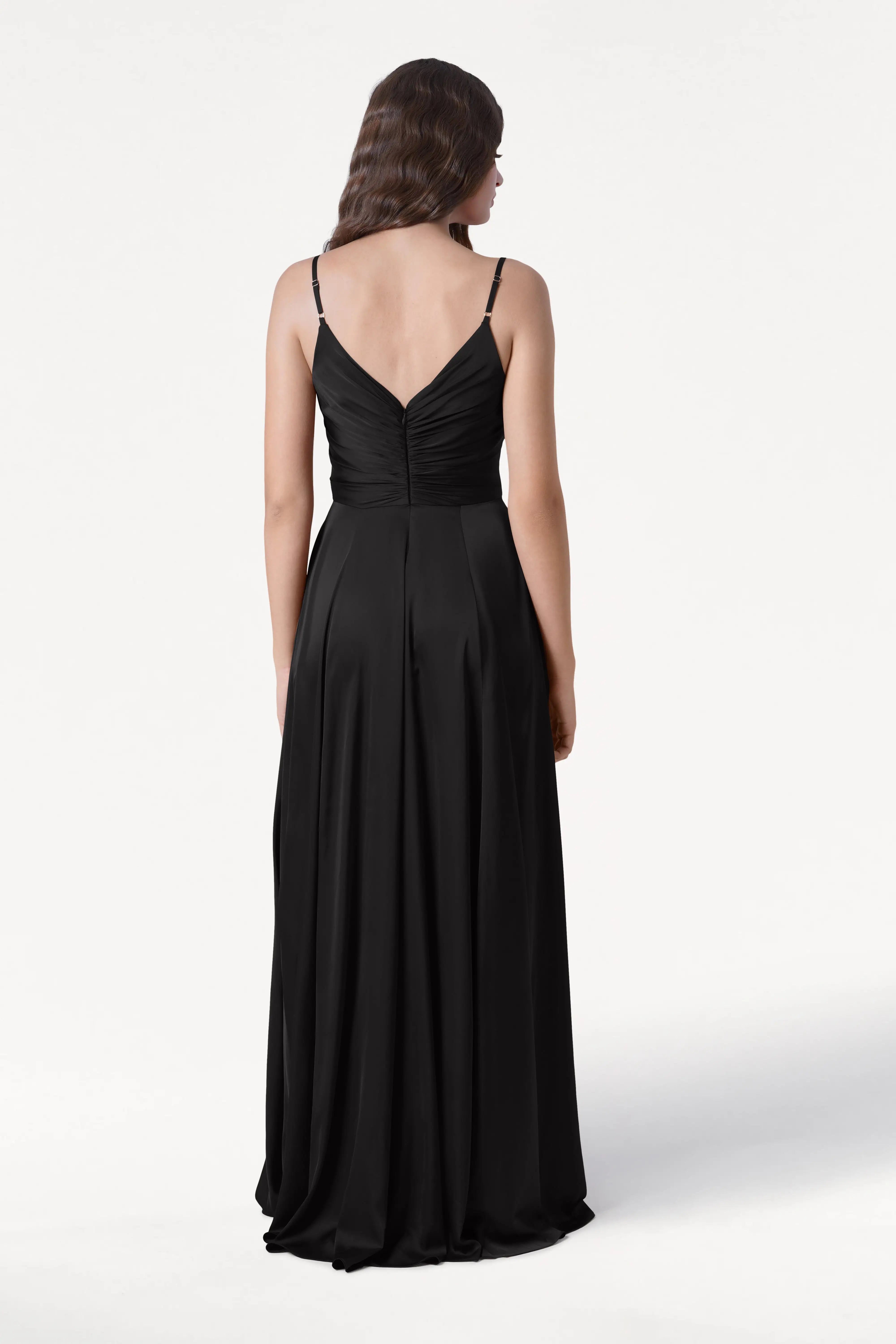 A woman with long, wavy brown hair is shown from the back wearing the TH&TH Isla Bridesmaid Dress in Black Satin, featuring thin straps and a pleated bodice. The background is plain white.
