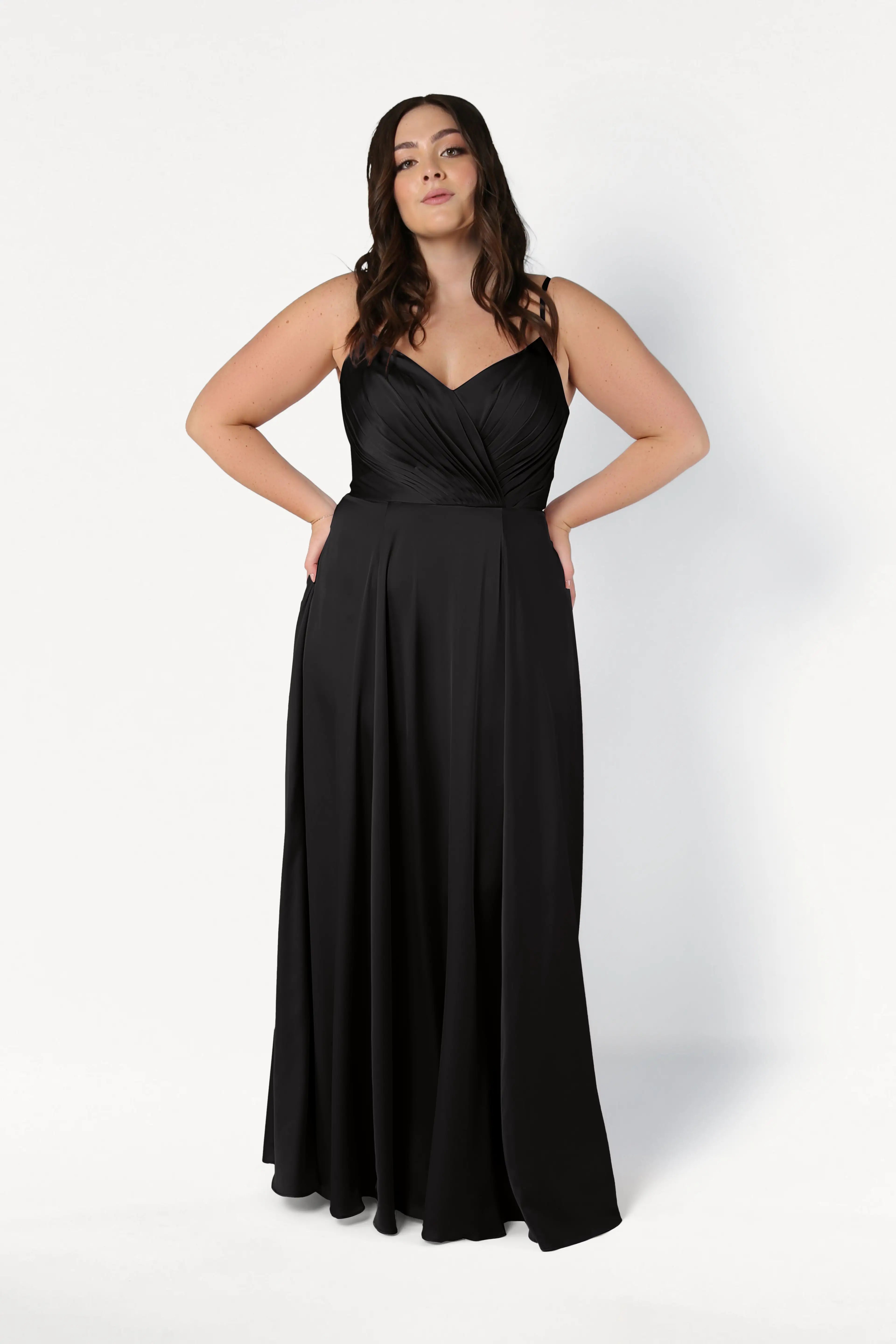 A woman stands against a plain white background, confidently posing with her hands on her hips in the TH&TH Isla Bridesmaid Dress in Black Satin, featuring a fitted bodice and flowing skirt.