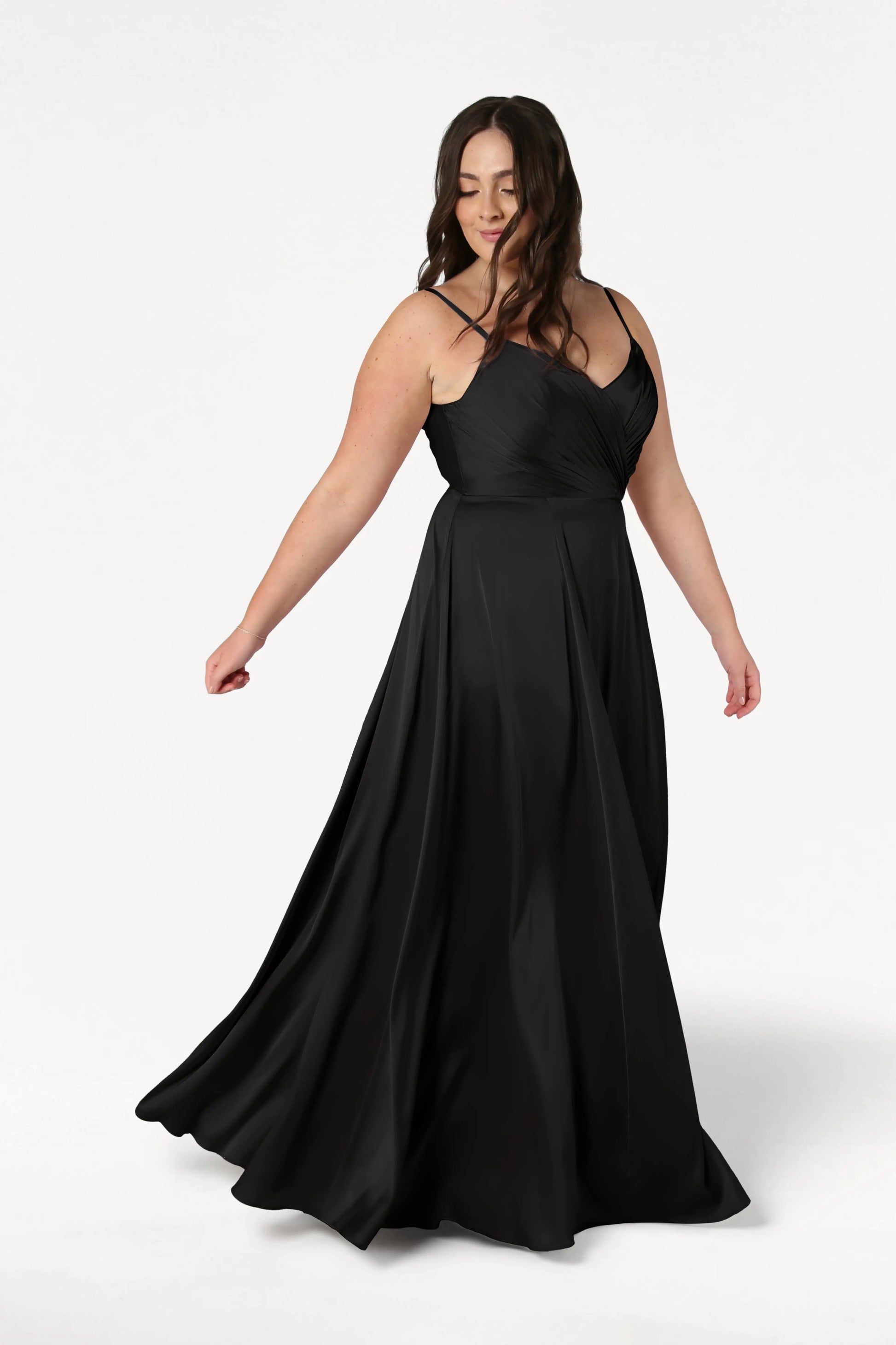 A woman with long dark hair wears the TH&TH Isla Bridesmaid Dress in Black Satin, its pleated bodice and floor-length skirt flowing elegantly as she stands against a white background, gracefully holding out the skirt.