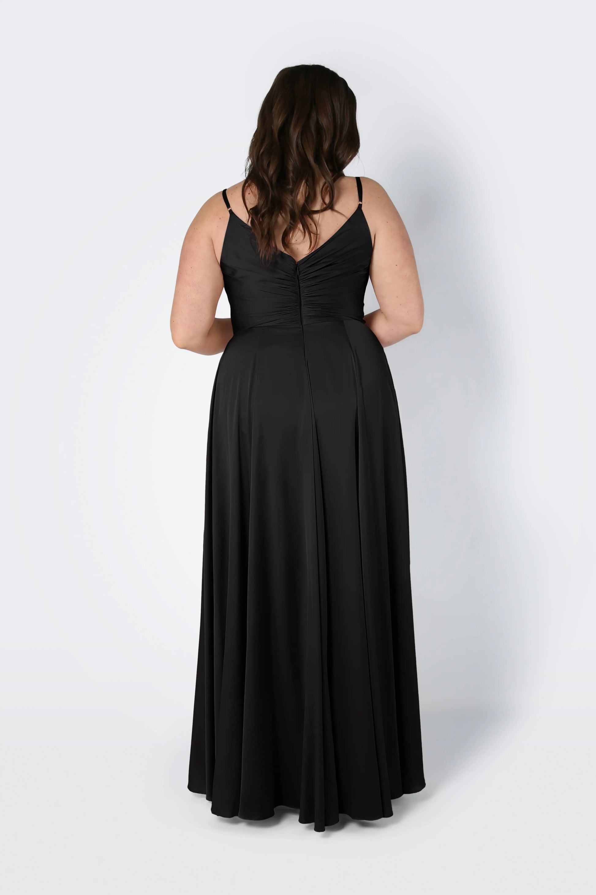A woman with long brown hair, shown from the back, wears the TH&TH Isla Bridesmaid Dress in black satin—floor-length, sleeveless with thin straps and a pleated bodice—standing against a plain white background.