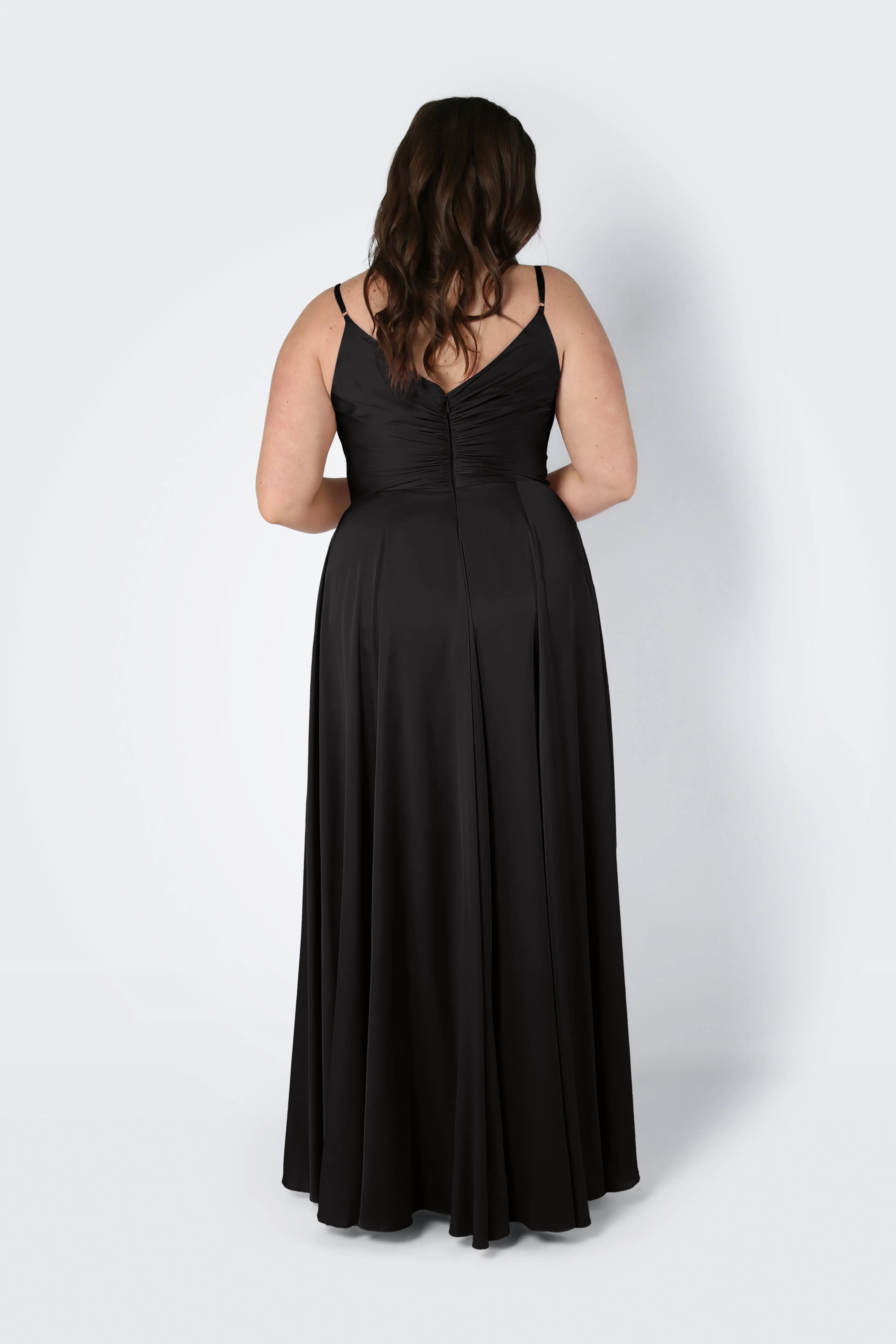 A woman with long brown hair, shown from the back, wears the TH&TH Isla Bridesmaid Dress in black satin—floor-length, sleeveless with thin straps and a pleated bodice—standing against a plain white background.