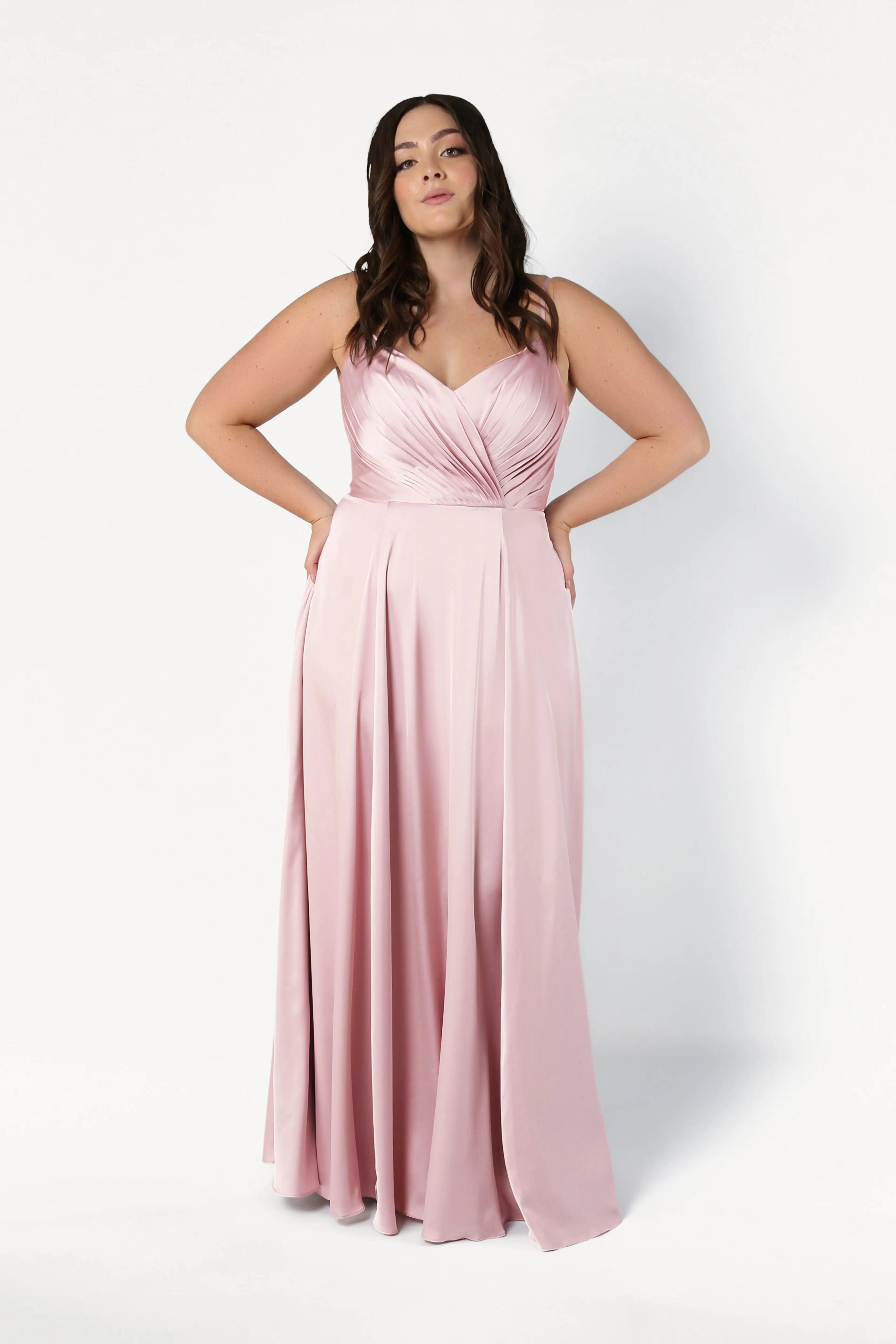 A woman wears the TH&TH Isla Bridesmaid Dress in blush pink satin, featuring a pleated bodice and spaghetti straps, as she stands confidently against a white background with her hands at her hips.