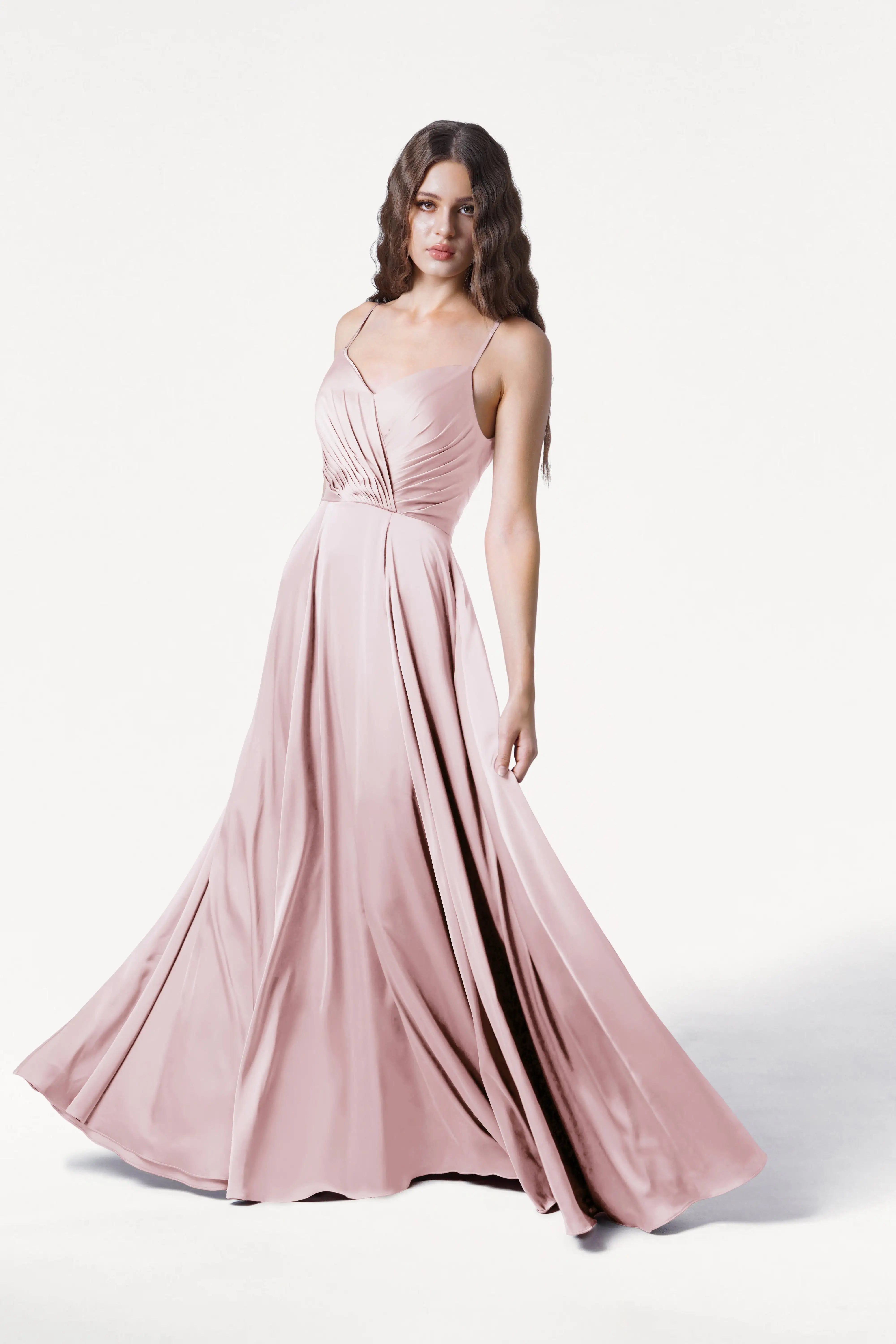 A woman in the TH&TH Isla Bridesmaid Dress in Blush Pink Satin, featuring thin straps and a pleated bodice, stands against a white background, gently holding the skirt as it fans out around her.