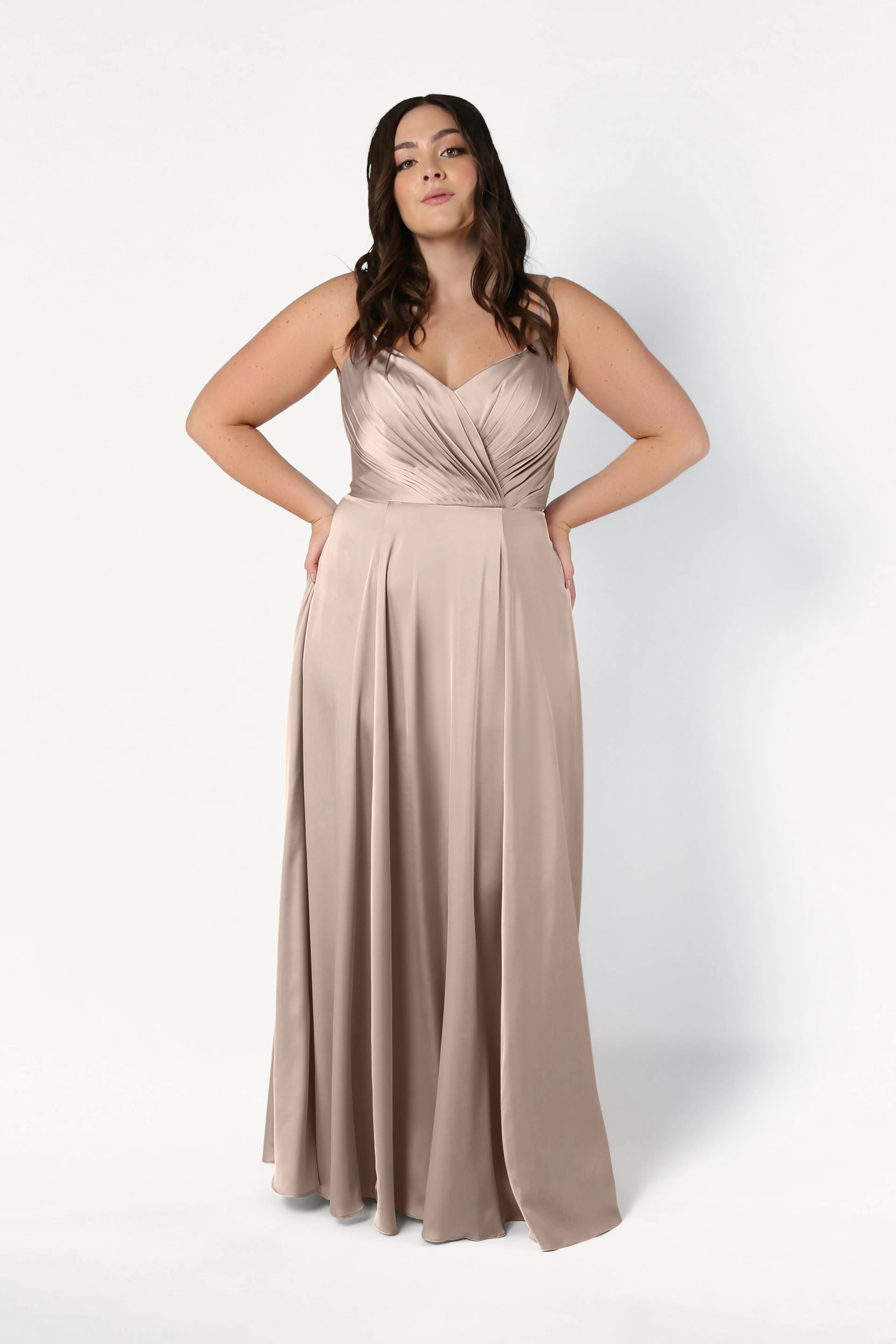 A plus-size model wears the Isla Bridesmaid Dress in Champagne Sky Satin by TH&TH, featuring a pleated bodice, spaghetti straps, and a flowing A-line skirt, posing with hands on hips against a white background.