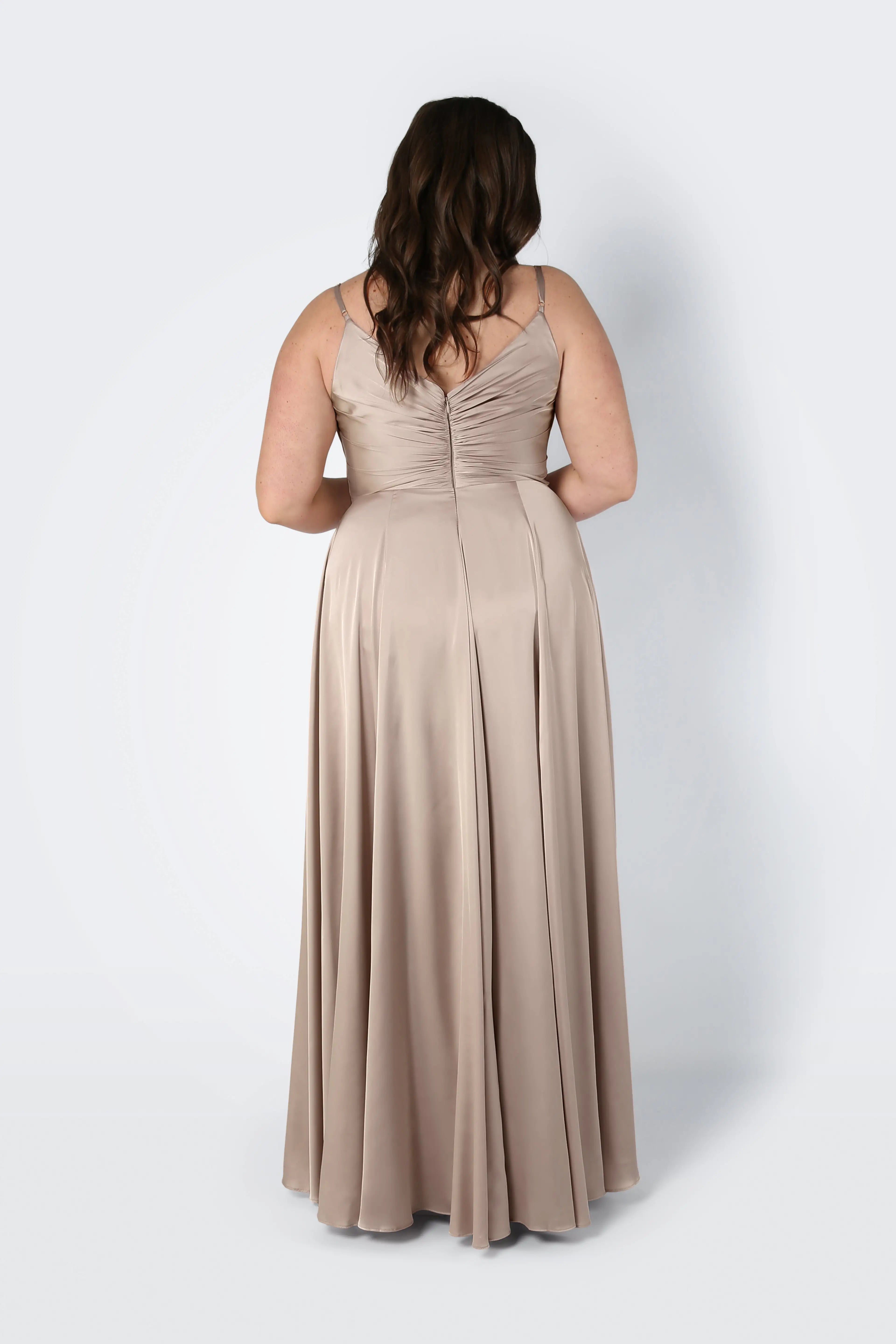 A woman with wavy brown hair is seen from behind in the TH&TH Isla Bridesmaid Dress in Champagne Sky Satin, featuring thin straps, a pleated bodice, and ruched back, standing against a plain light background.