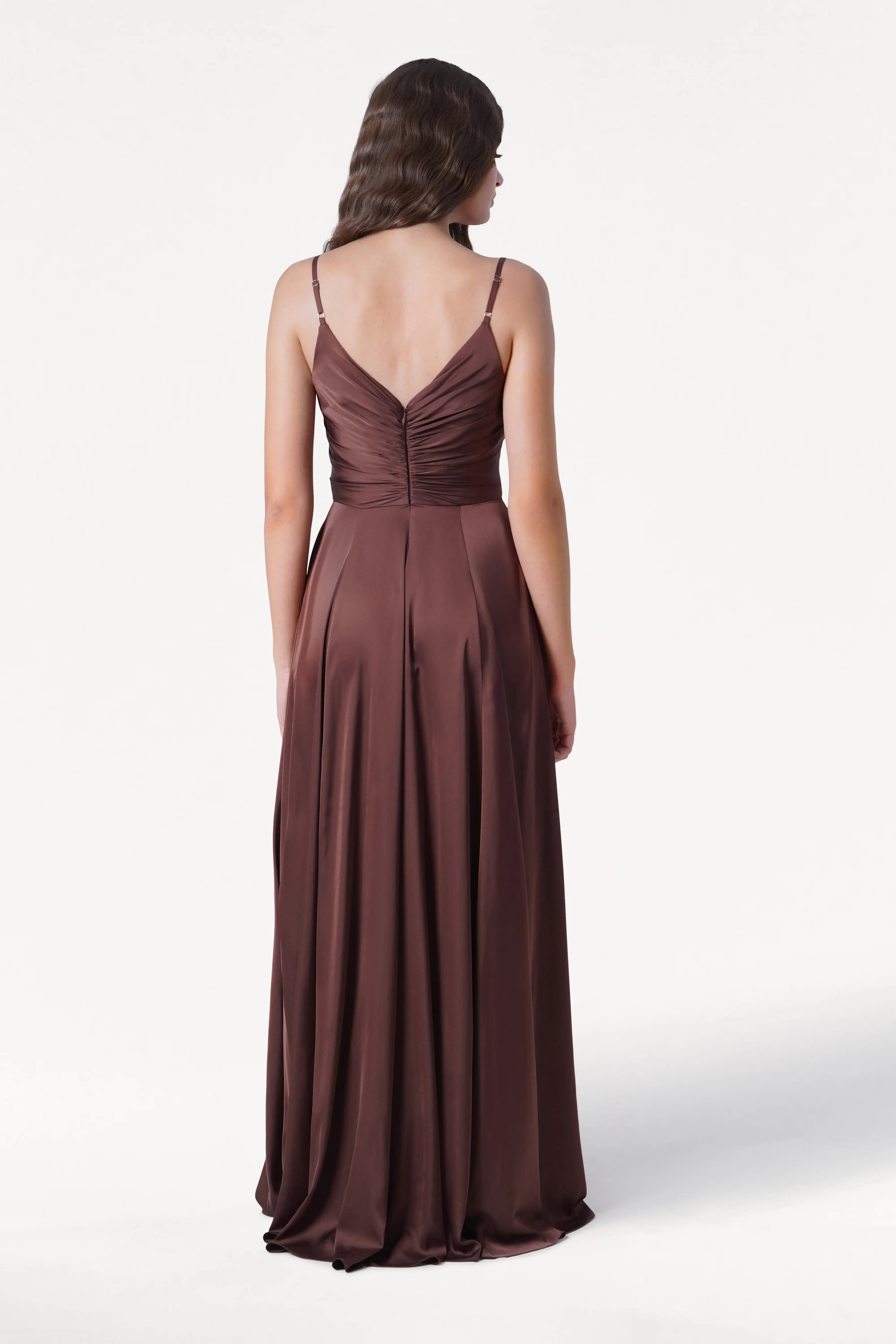A woman with wavy brown hair faces away, wearing the TH&TH Isla Bridesmaid Dress in chocolate brown satin. The gown features thin straps, a pleated bodice, and flows smoothly to the floor against a plain white background.