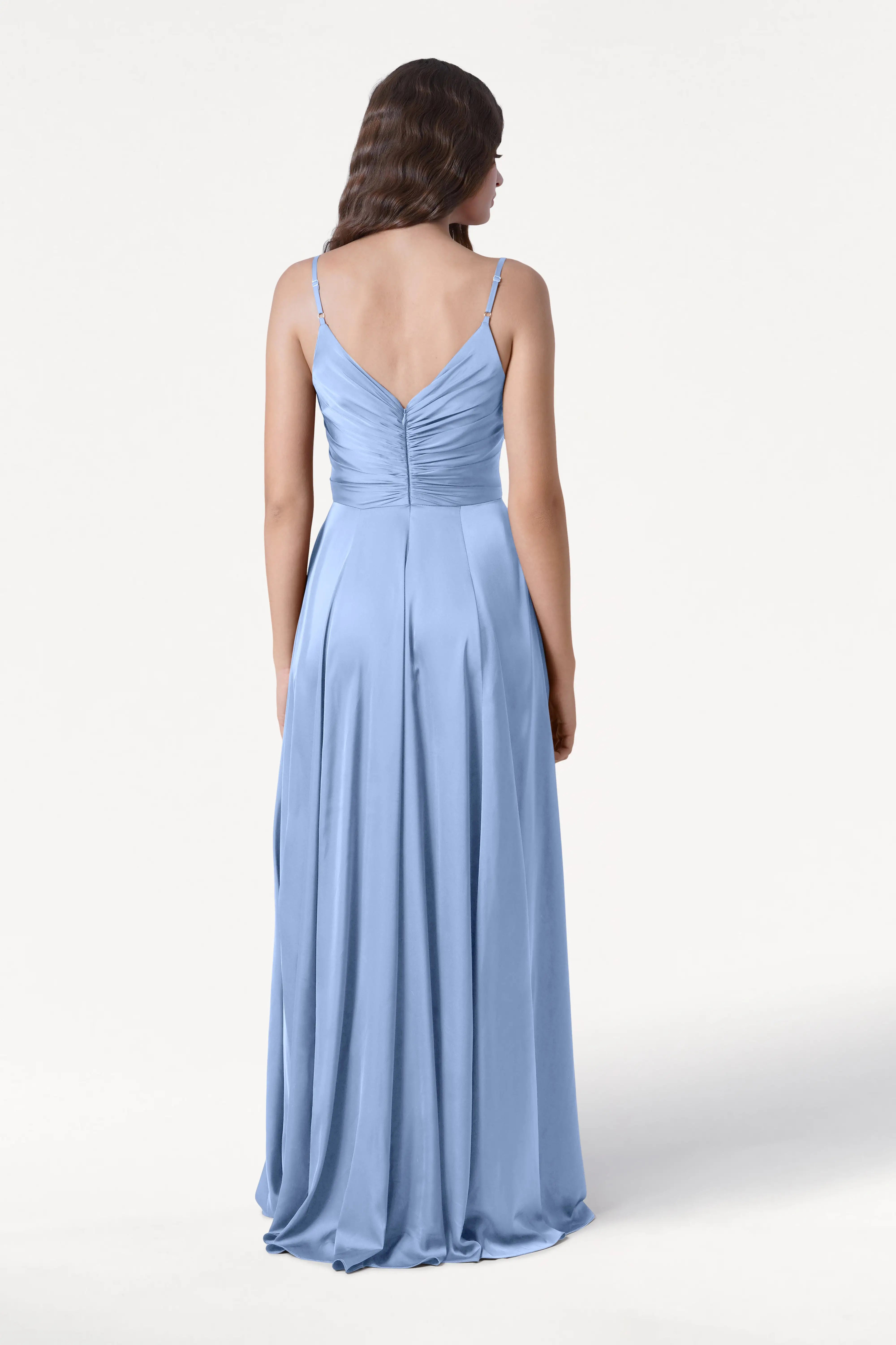 A woman faces away wearing the TH&TH Isla Bridesmaid Dress in Dusty Blue Satin—a floor-length gown with thin straps and a pleated bodice. Her wavy brown hair cascades down her back against a plain white background.