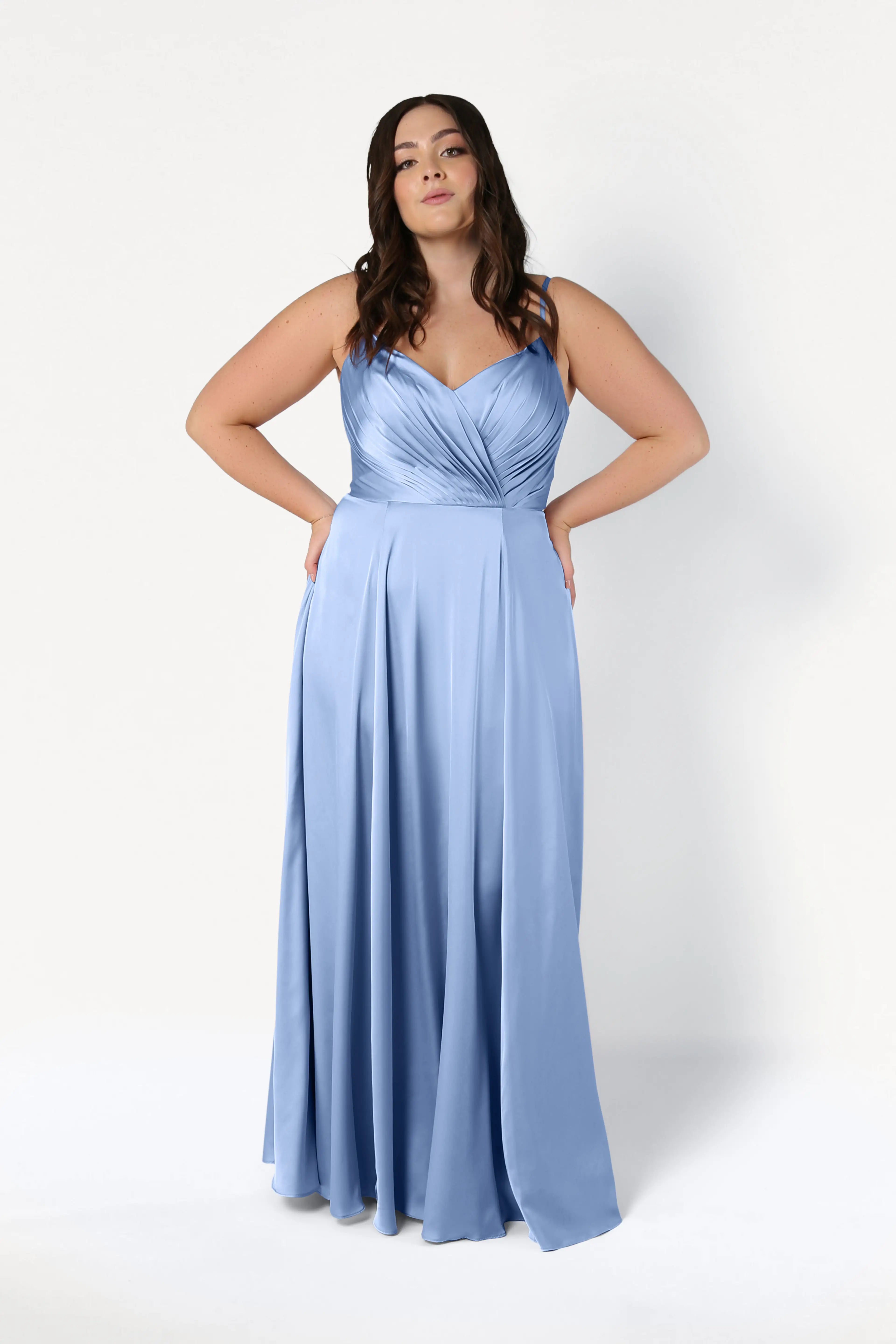 A woman poses in the Isla Bridesmaid Dress in Dusty Blue Satin by TH&TH—a long satin dress with thin straps and a pleated bodice—against a plain white background.