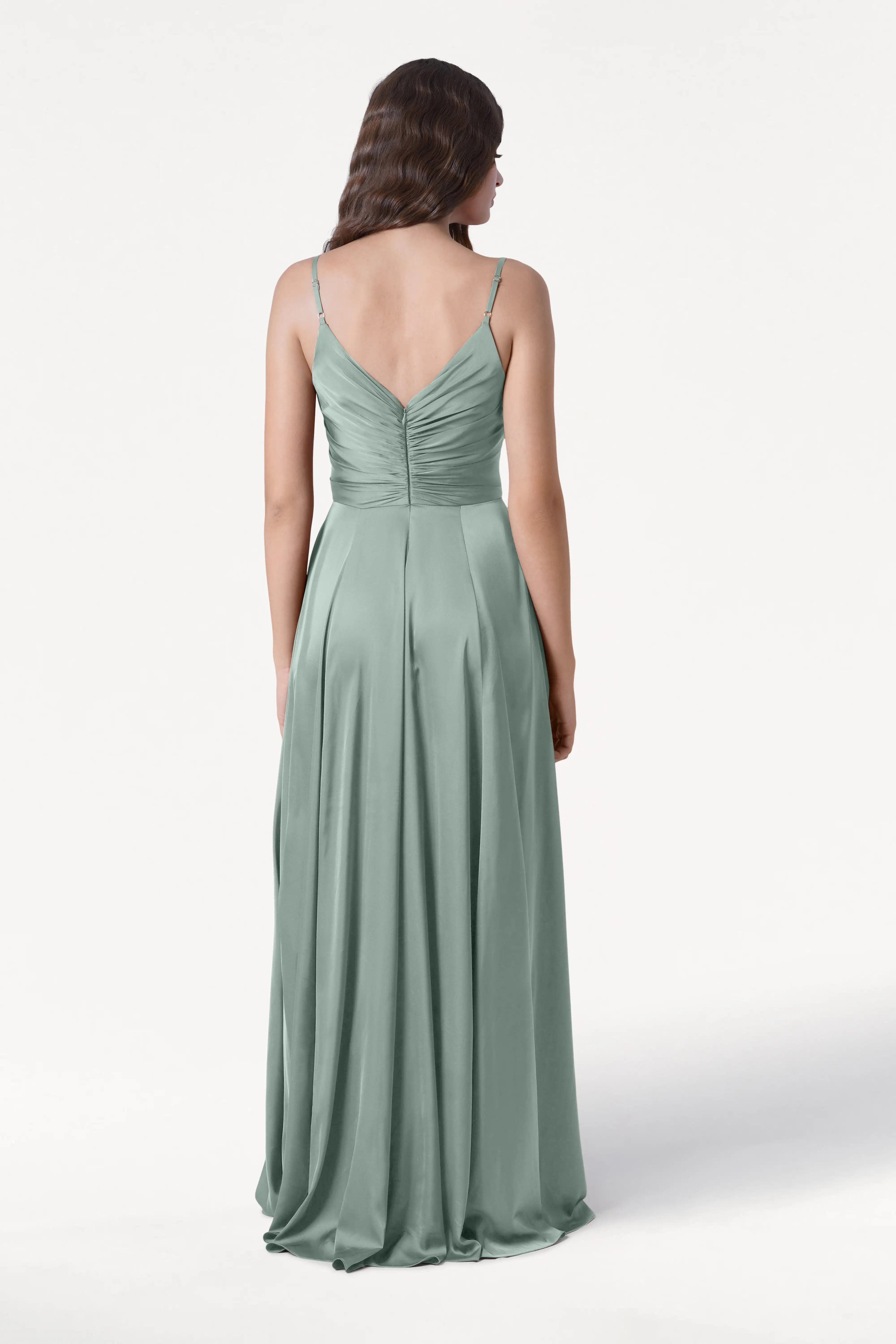 A woman with wavy brown hair wears the TH&TH Isla Bridesmaid Dress in Dusty Eucalyptus Satin, featuring thin straps, a pleated bodice, and a gathered V-shaped back. She stands facing away from the camera against a plain white background.
