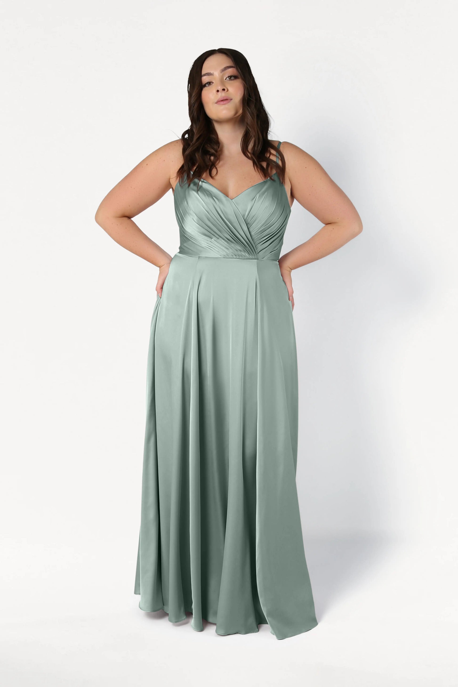 A woman with long brown hair wears the TH&TH Isla Bridesmaid Dress in Dusty Eucalyptus Satin, featuring a pleated bodice and flowing skirt. She stands against a plain white background with her hands on her hips.