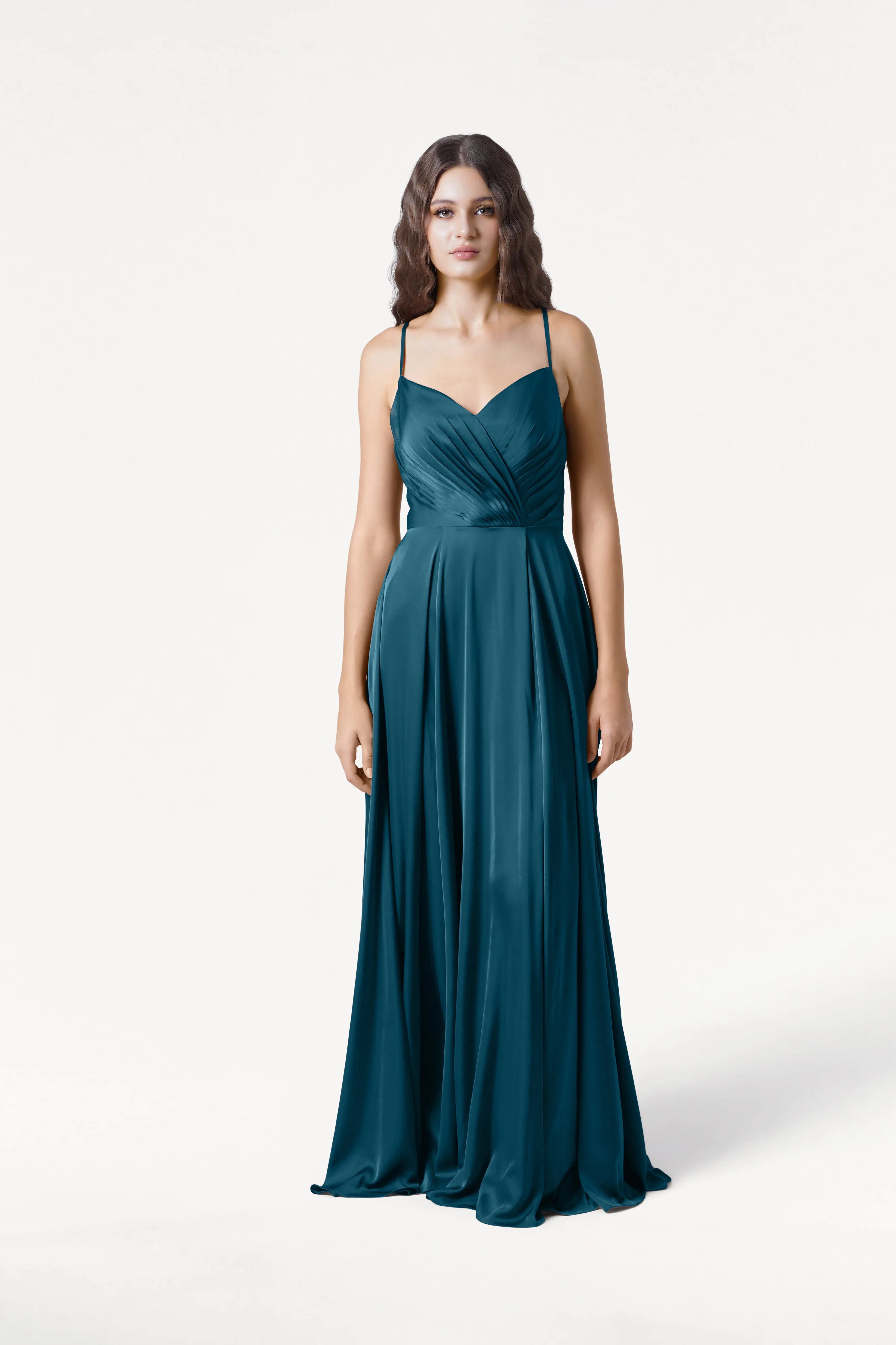 A woman with wavy brown hair wears the TH&TH Isla Bridesmaid Dress in Emerald Dawn Satin, a floor-length gown with spaghetti straps and a pleated bodice, standing against a plain white background.