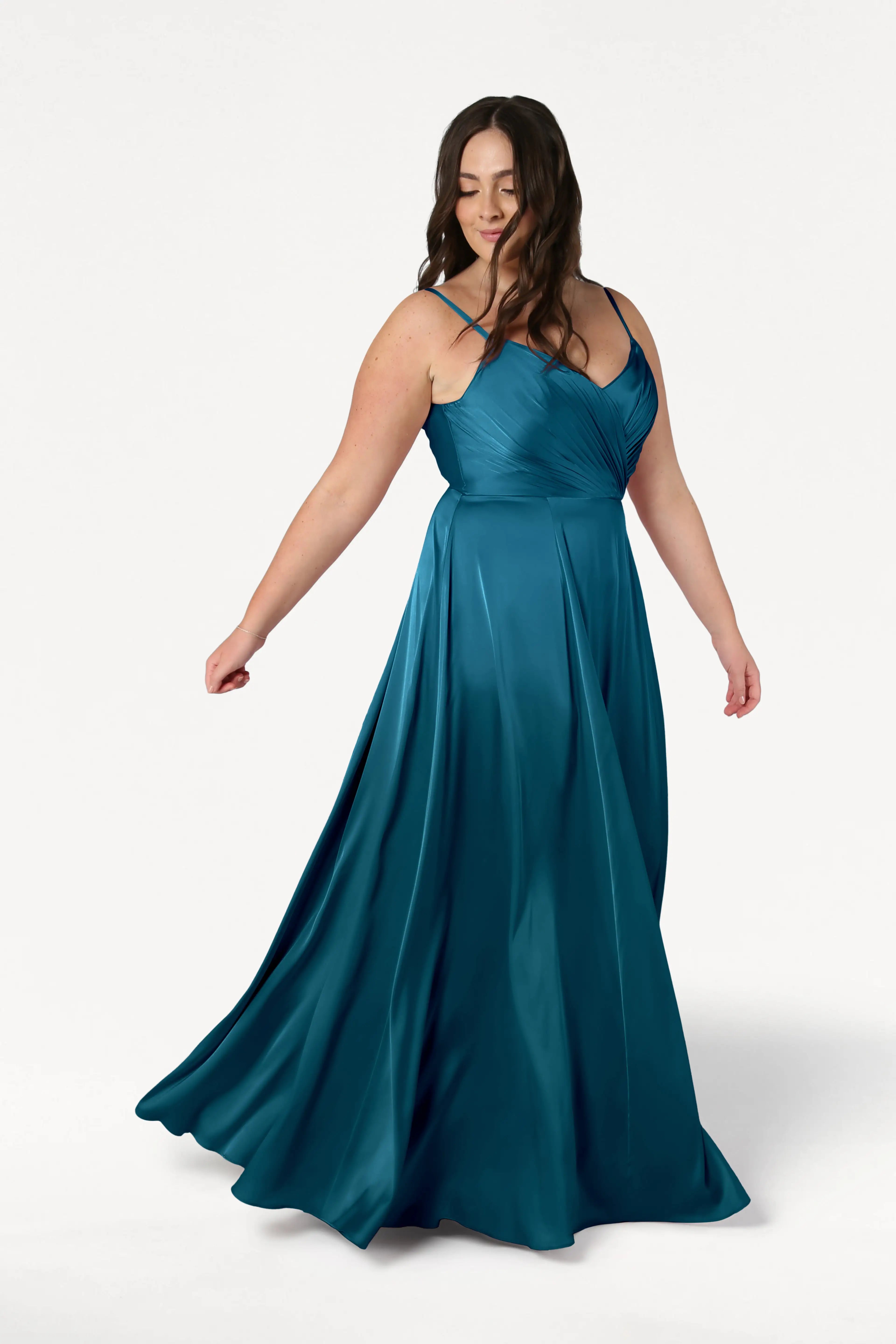 A woman with long, wavy brown hair twirls gracefully in the Isla Bridesmaid Dress in Emerald Dawn Satin by TH&TH, showing off its floor-length teal hue and pleated bodice against a plain white background.