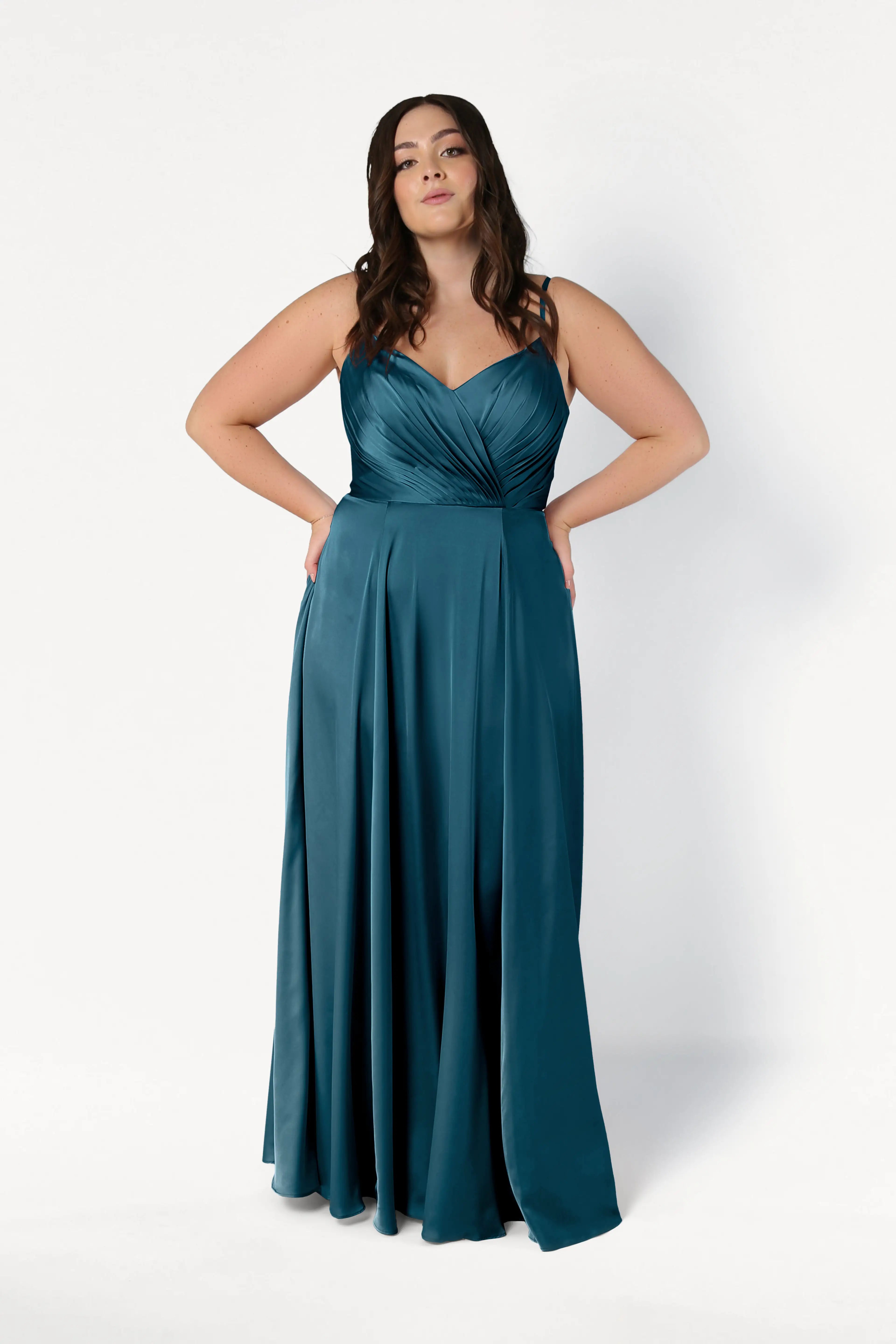 A woman stands against a white background wearing the TH&TH Isla Bridesmaid Dress in Emerald Dawn Satin, a long gown with a pleated bodice and flowing skirt. Her hands are on her hips, and her dark hair falls in loose waves.