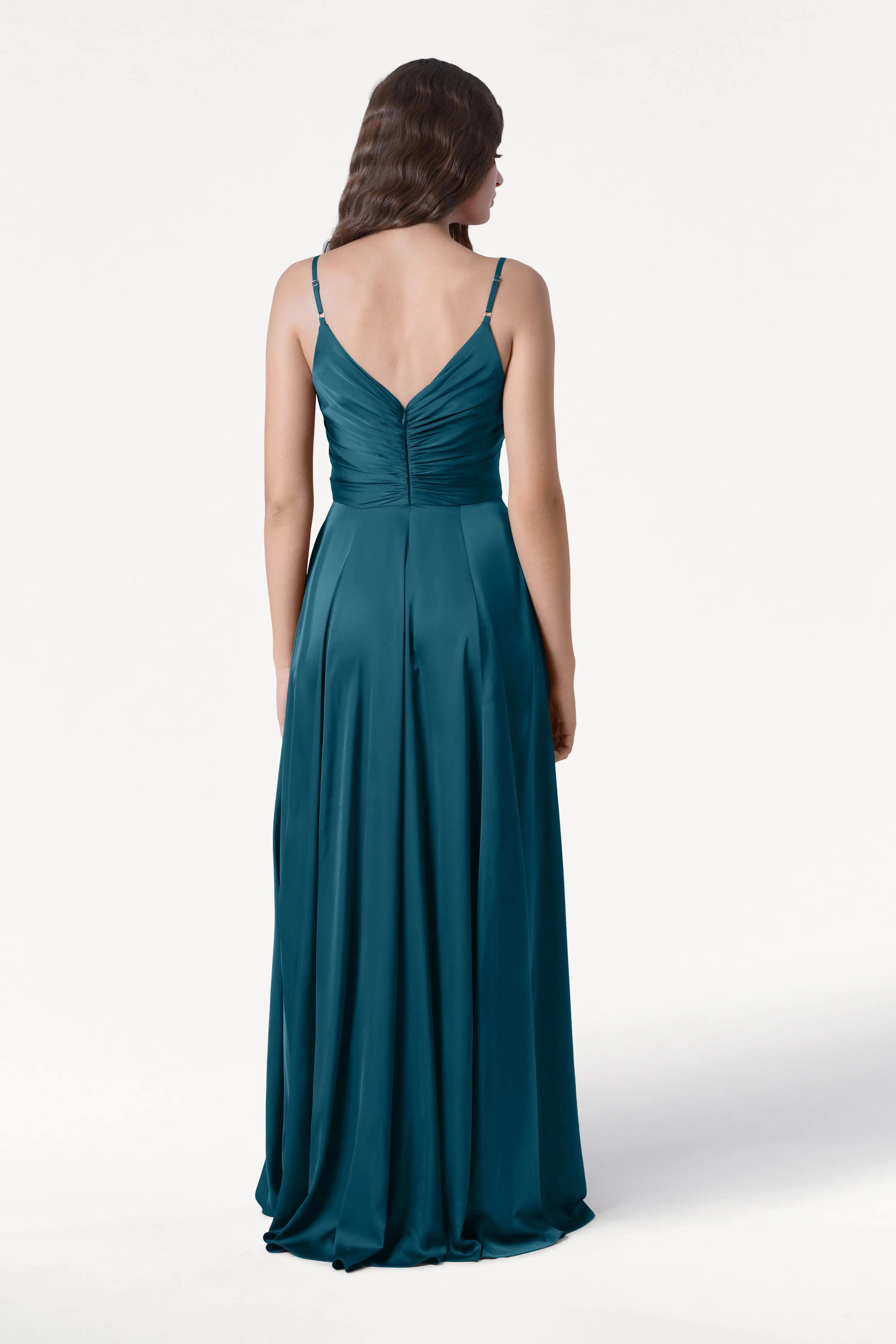 A woman with long wavy brown hair stands facing away, wearing the TH&TH Isla Bridesmaid Dress in Emerald Dawn Satin—a floor-length gown with thin straps, a pleated bodice, and a V-shaped back against a white background.