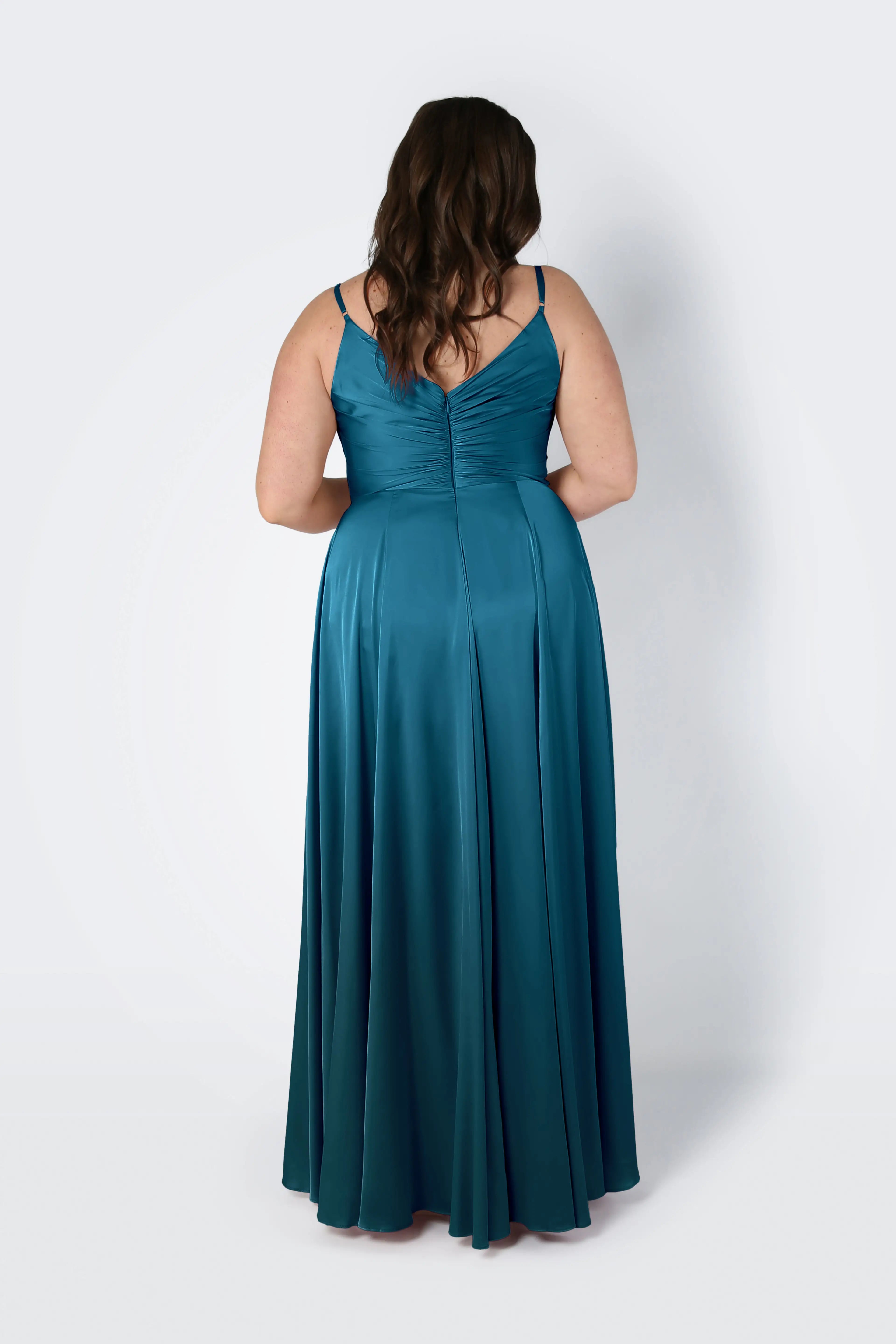 A woman with long brown hair stands with her back to the camera, wearing the Isla Bridesmaid Dress in Emerald Dawn Satin by TH&TH—a floor-length gown with a pleated bodice and spaghetti straps, set against a plain light background.