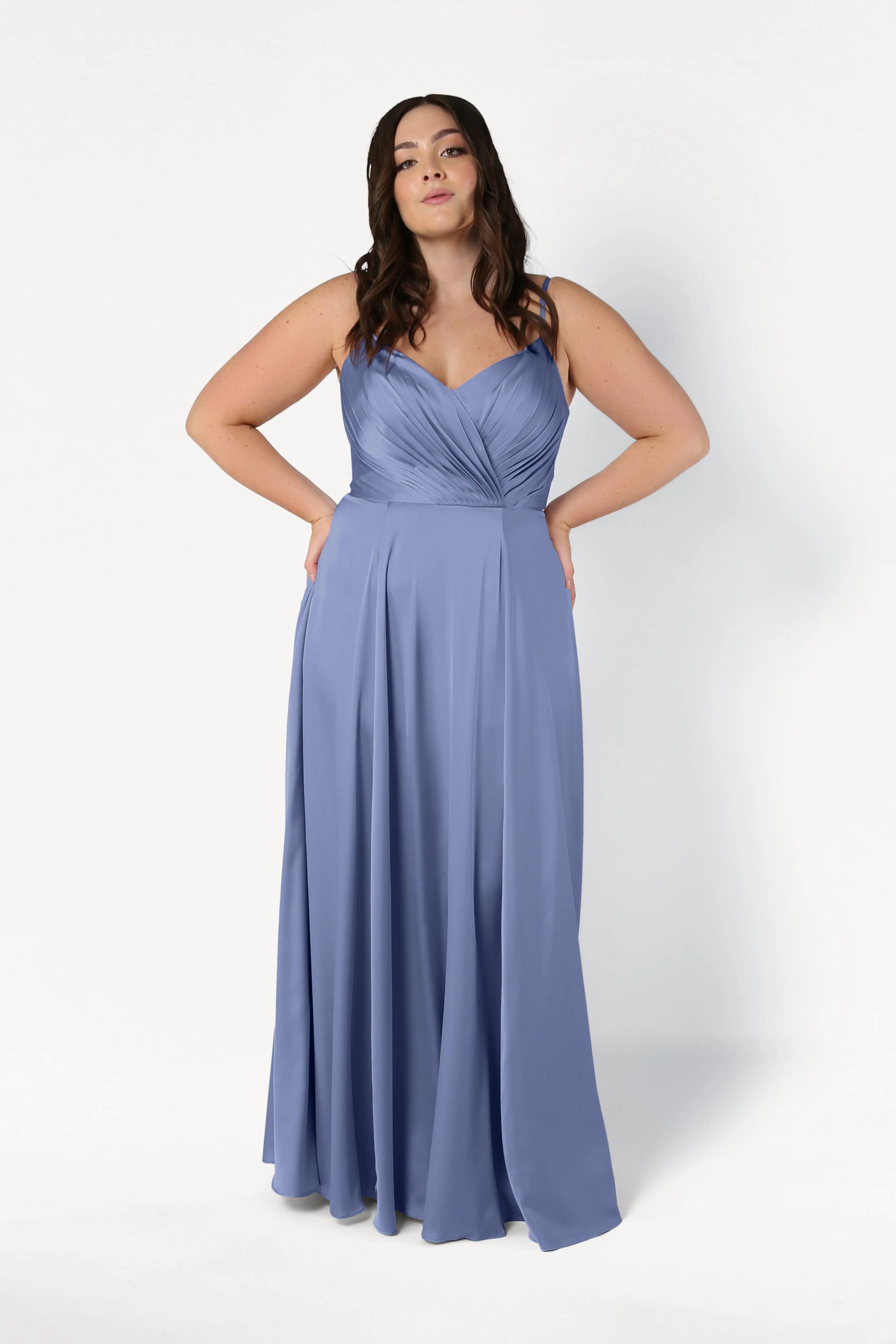 A woman with long, wavy dark hair stands against a white background wearing the TH&TH Isla Bridesmaid Dress in French Blue Satin, featuring spaghetti straps and a pleated bodice. Her hands rest on her hips.