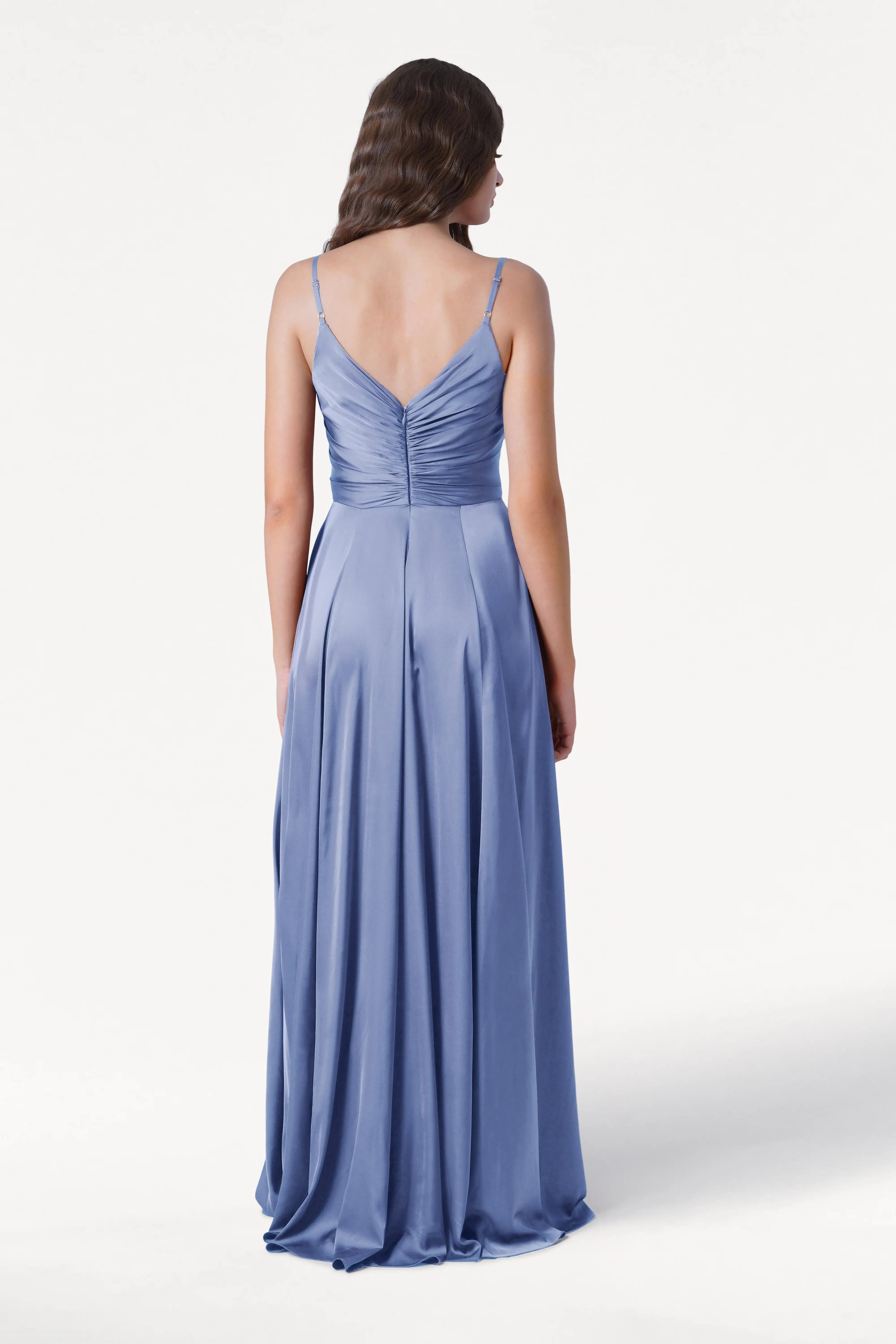 A woman with wavy brown hair stands with her back to the camera, wearing the TH&TH Isla Bridesmaid Dress in French Blue Satin—an elegant long dress with thin straps and a pleated bodice—set against a plain white background.