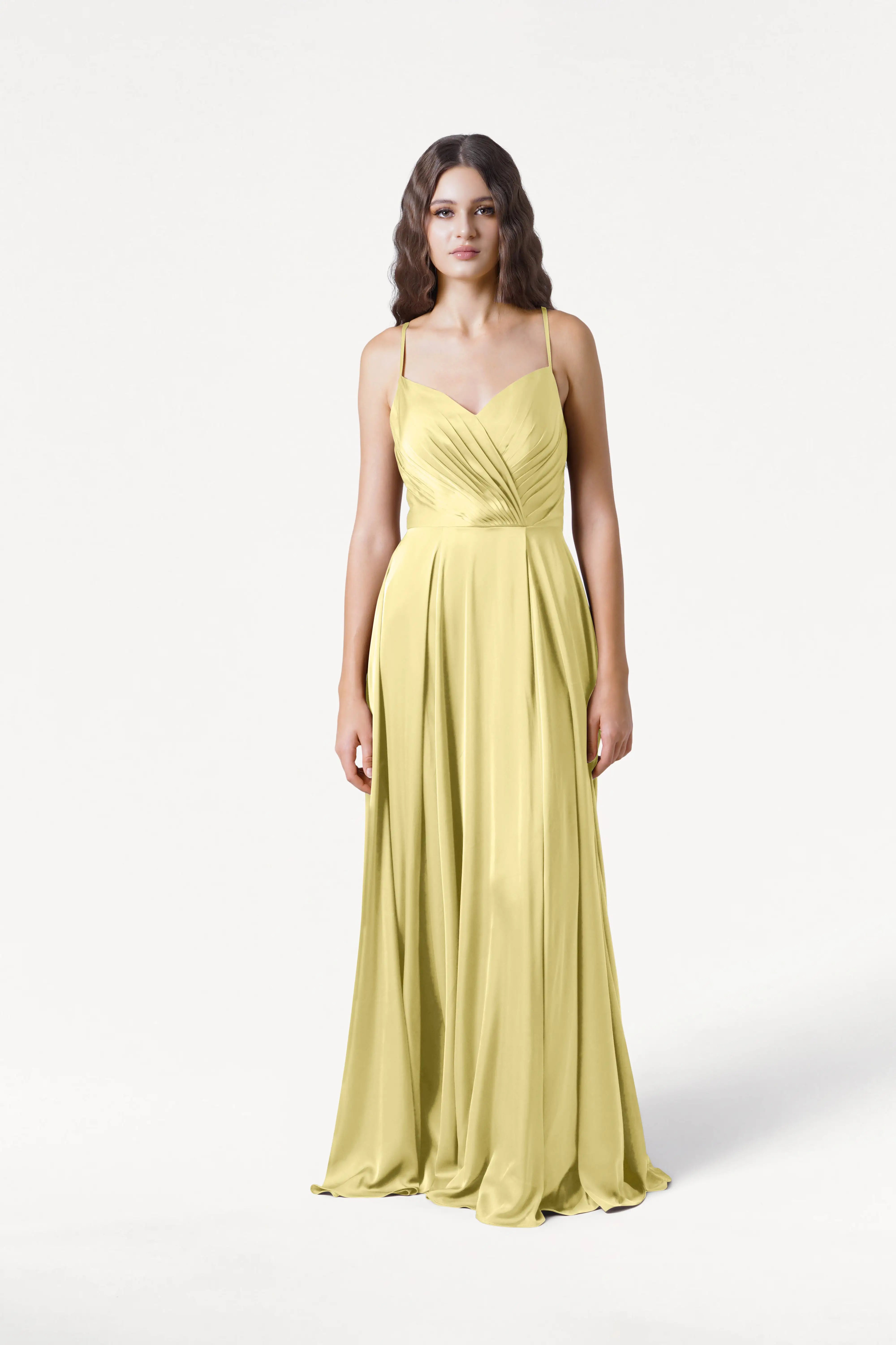 A woman with long, wavy hair and a neutral expression stands against a white background wearing the TH&TH Isla Bridesmaid Dress in Limoncello Satin, a long yellow gown with thin straps and a fitted, pleated bodice.