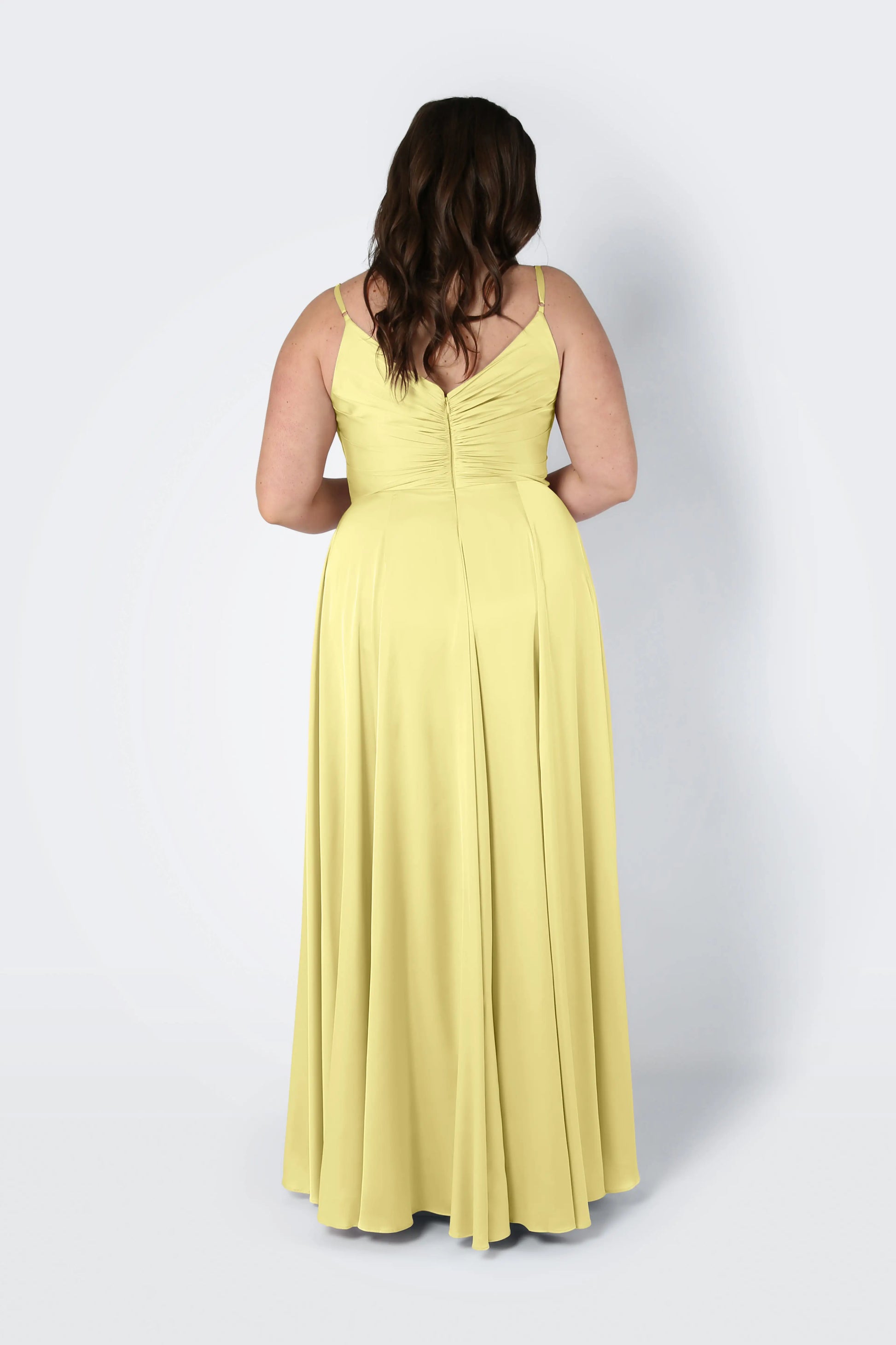A woman with wavy brown hair faces away, wearing the Isla Bridesmaid Dress in Limoncello Satin by TH&TH—a long, flowing yellow satin dress with thin straps and a pleated bodice—set against a plain light background.