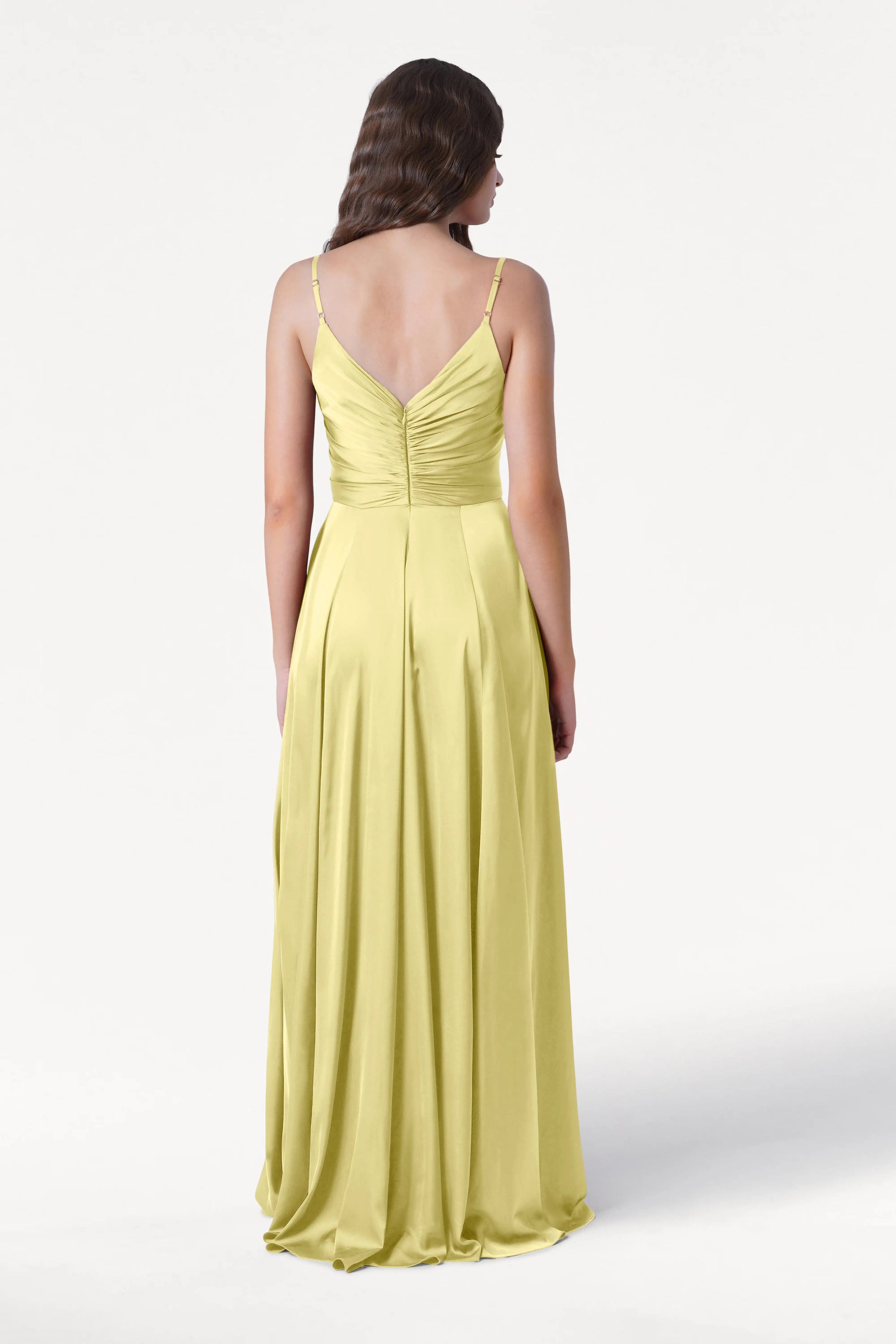 A woman with loose brown waves stands against a white background, wearing the TH&TH Isla Bridesmaid Dress in Limoncello Satin—a long satin gown with thin straps, pleated bodice, ruched back, and flowing skirt.