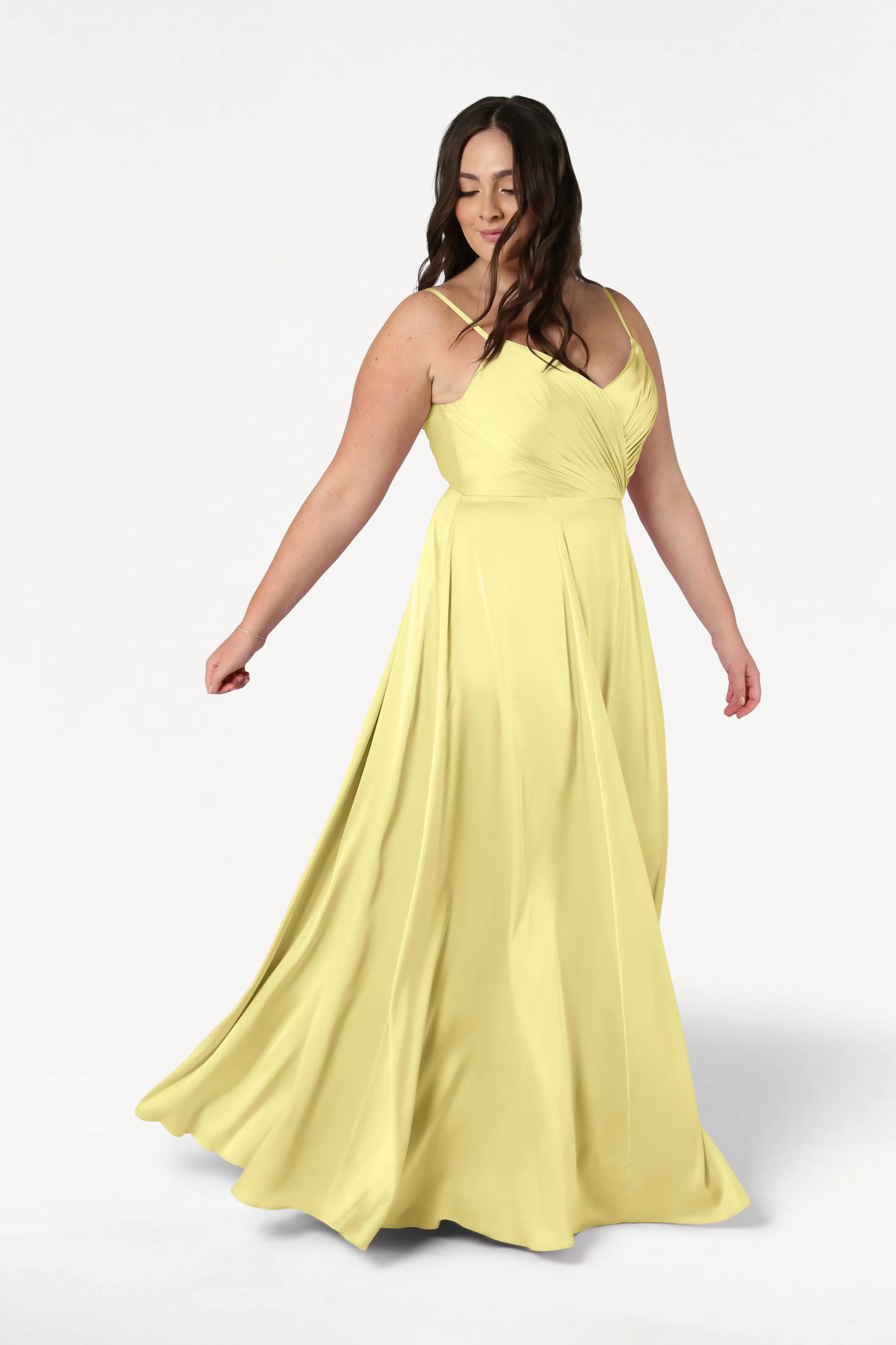 A woman twirls gracefully in the TH&TH Isla Bridesmaid Dress in Limoncello Satin, featuring a pleated bodice and flowing skirt, set against a plain white background.
