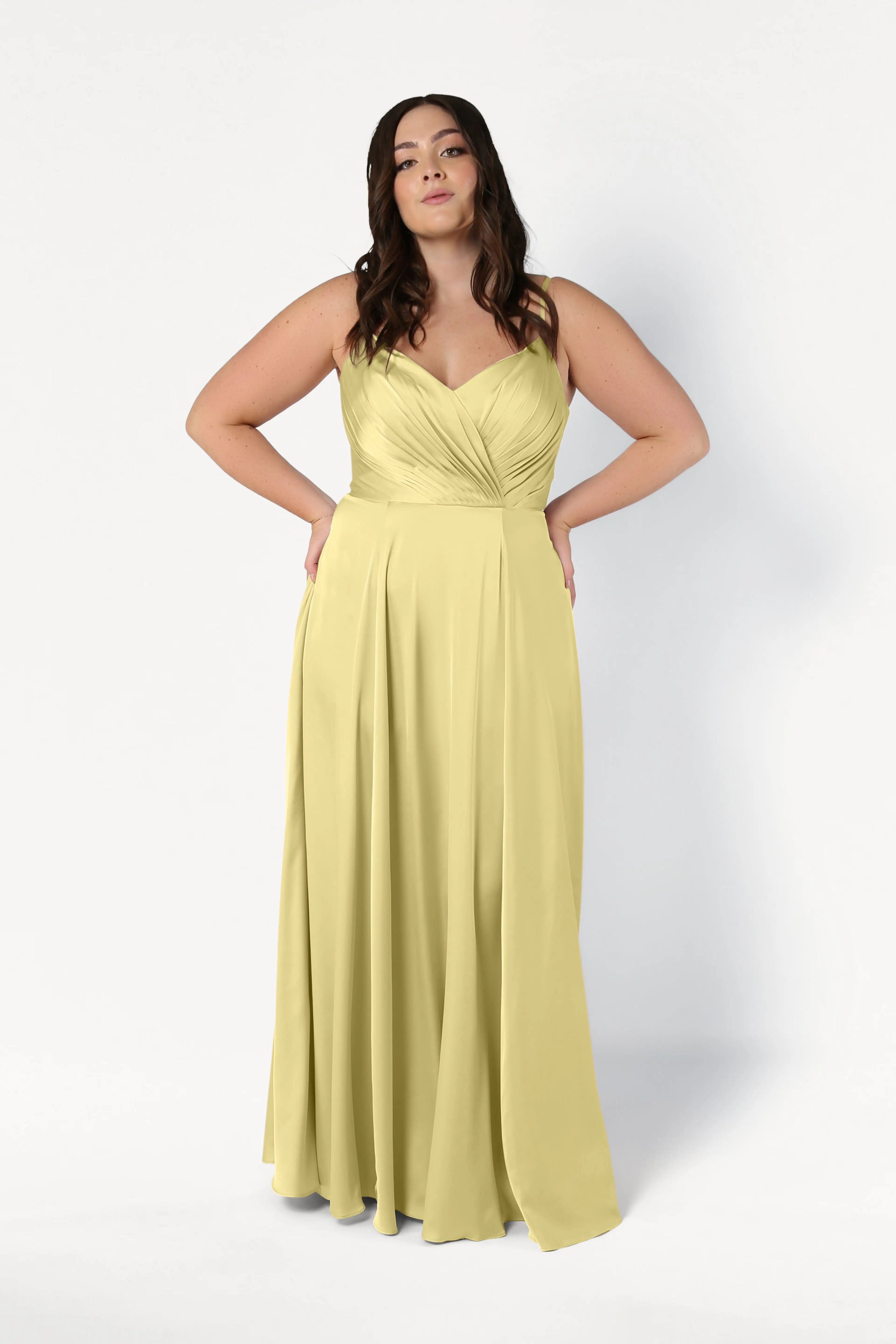 A woman models the TH&TH Isla Bridesmaid Dress in Limoncello Satin, a light yellow, floor-length gown with a pleated bodice, standing confidently against a plain white background with her hands on her hips.