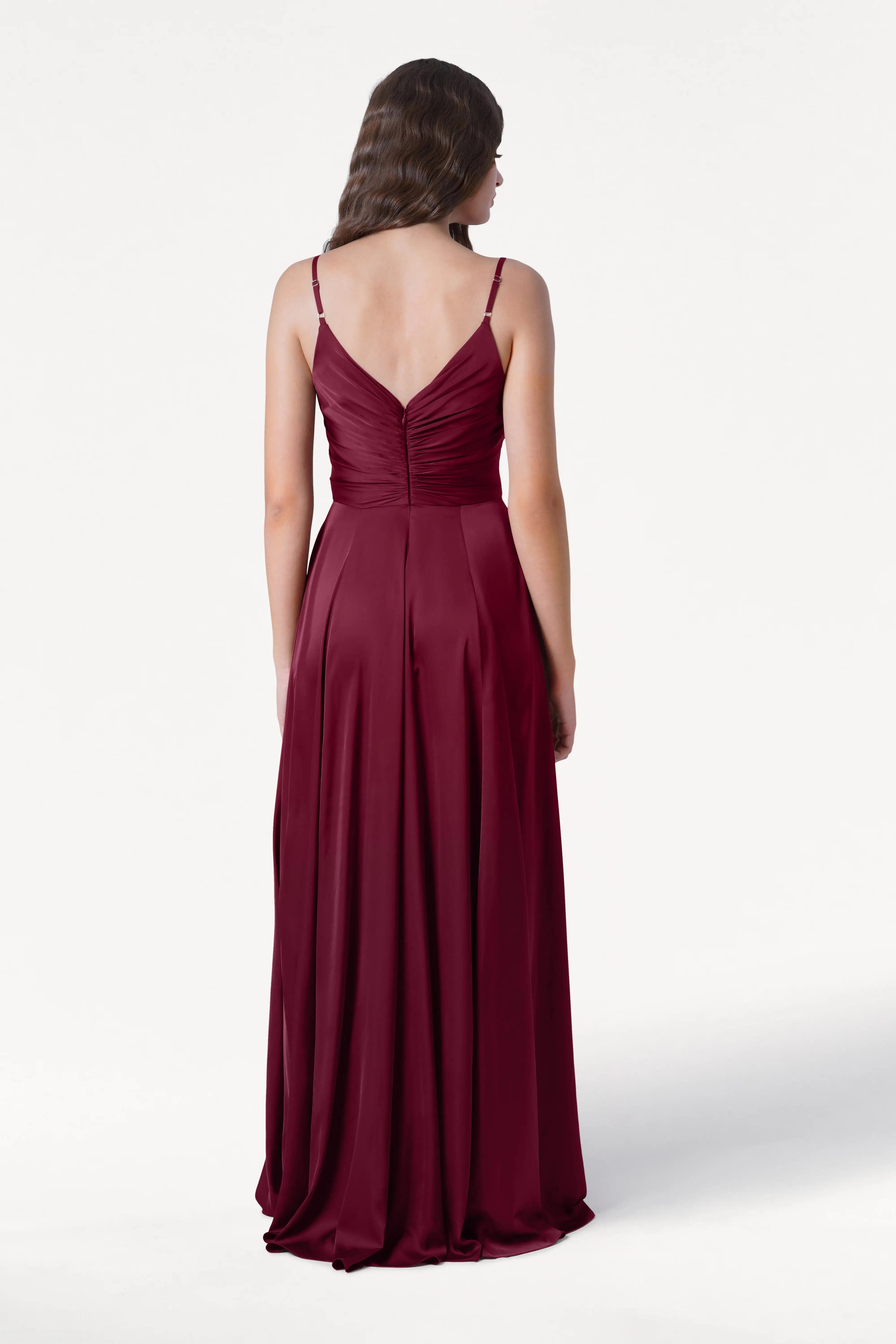 A woman with loose waves stands facing away against a white background, wearing the TH&TH Isla Bridesmaid Dress in Merlot Satin—a floor-length, sleeveless dress with thin straps and a fitted, slightly pleated bodice.